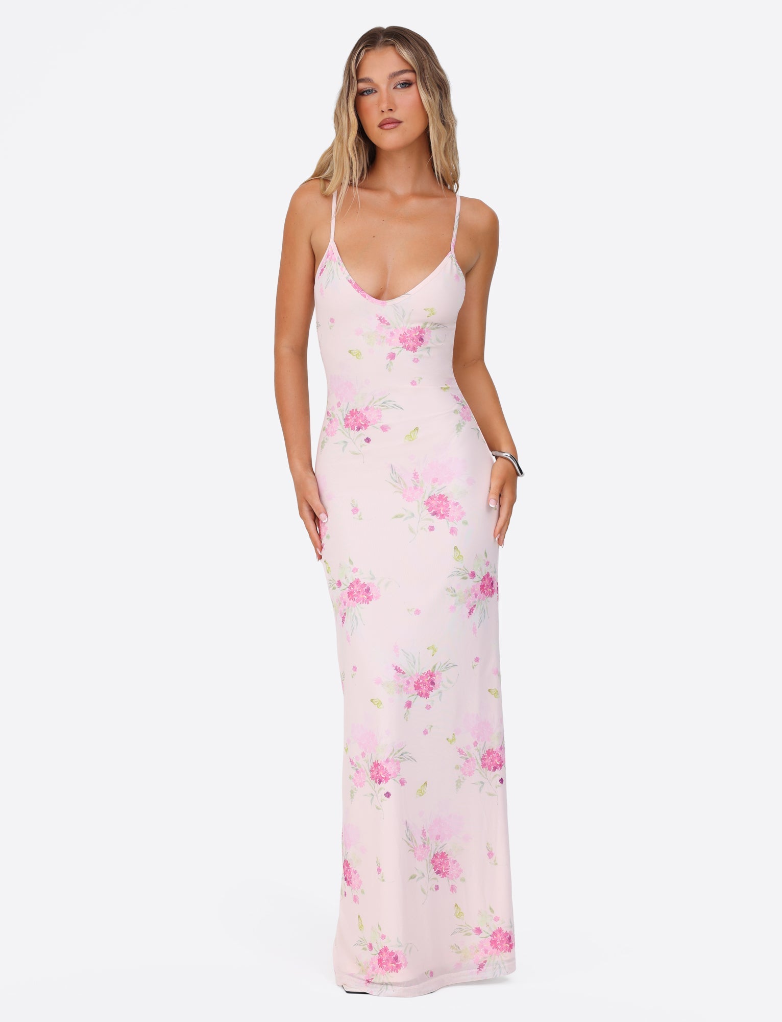 A woman wearing the OSIRIS DRESS - PINK : FLORAL : WATER COLOUR FLORAL by TIGERMIST stands against a plain white background. The maxi-length, sleeveless dress showcases intricate pink and green floral patterns. She stands with her hands by her sides, looking directly at the camera.
