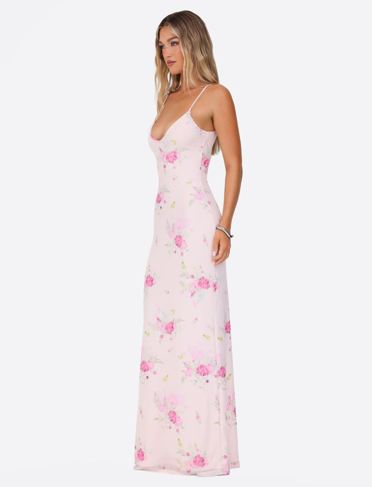 A woman with long blonde hair is wearing the OSIRIS DRESS - PINK : FLORAL : WATER COLOUR FLORAL by TIGERMIST. The dress features spaghetti straps and a delicate mesh fabrication, which highlights its elegant and flowing design as she stands in profile against a plain white background.