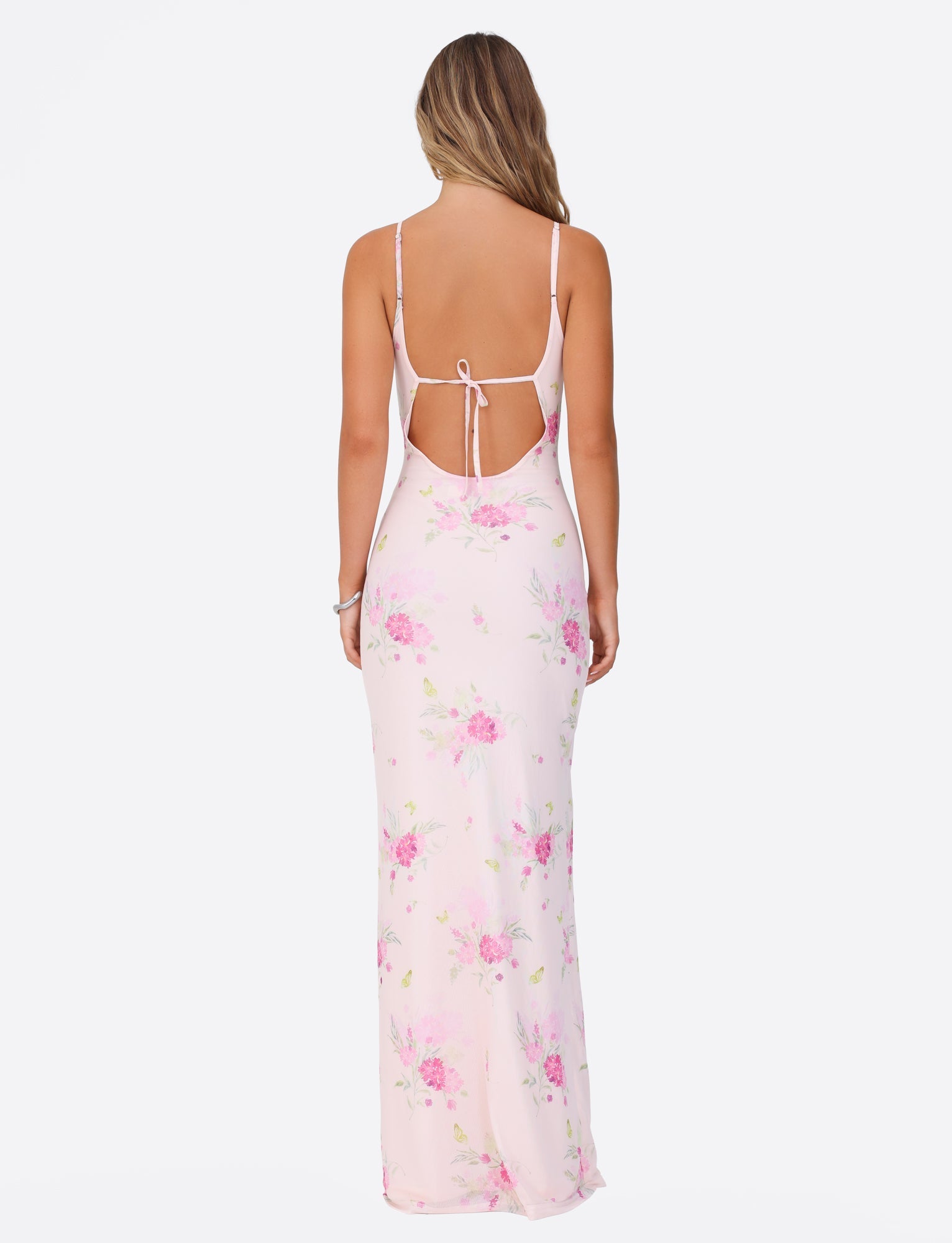 A woman showcases the TIGERMIST OSIRIS DRESS - PINK : FLORAL : WATER COLOUR FLORAL, as she stands facing away. The dress features a long maxi length and a backless design with thin shoulder straps and an open tie at the back, complemented by her long wavy hair. She is set against a plain white background.
