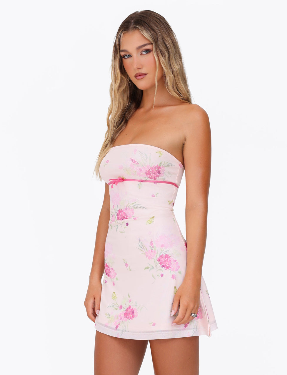 A woman with long blonde hair stands confidently against a plain white background, showcasing the elegant asymmetrical hemline of the CARSYN MINI DRESS by TIGERMIST, featuring a strapless design in pink with a watercolour floral print.