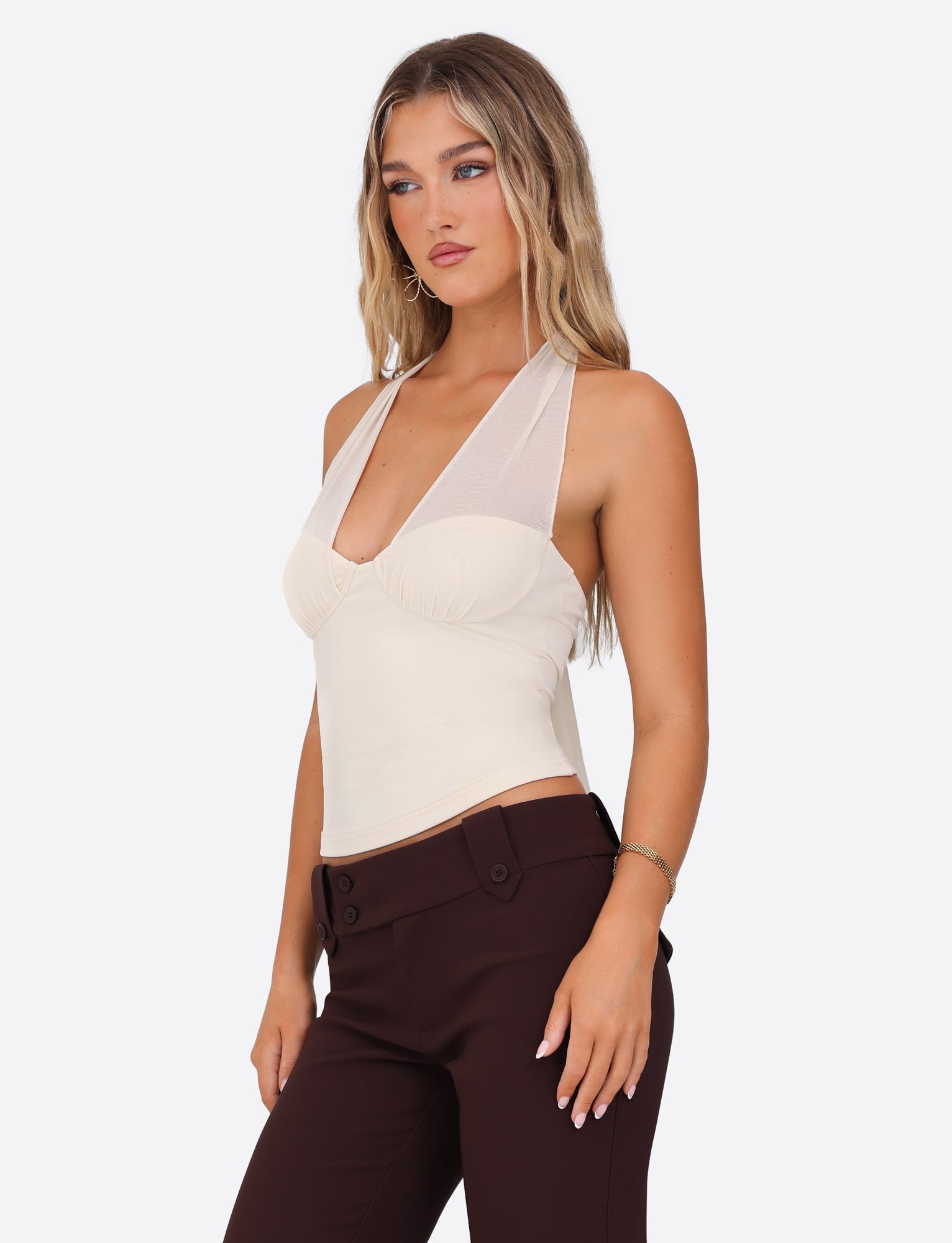 A young woman with long wavy hair is wearing a TIGERMIST GRANGER TOP in white cream, which features a halter neckline and mesh detail at the straps, paired with dark brown pants. She poses against a plain white background.