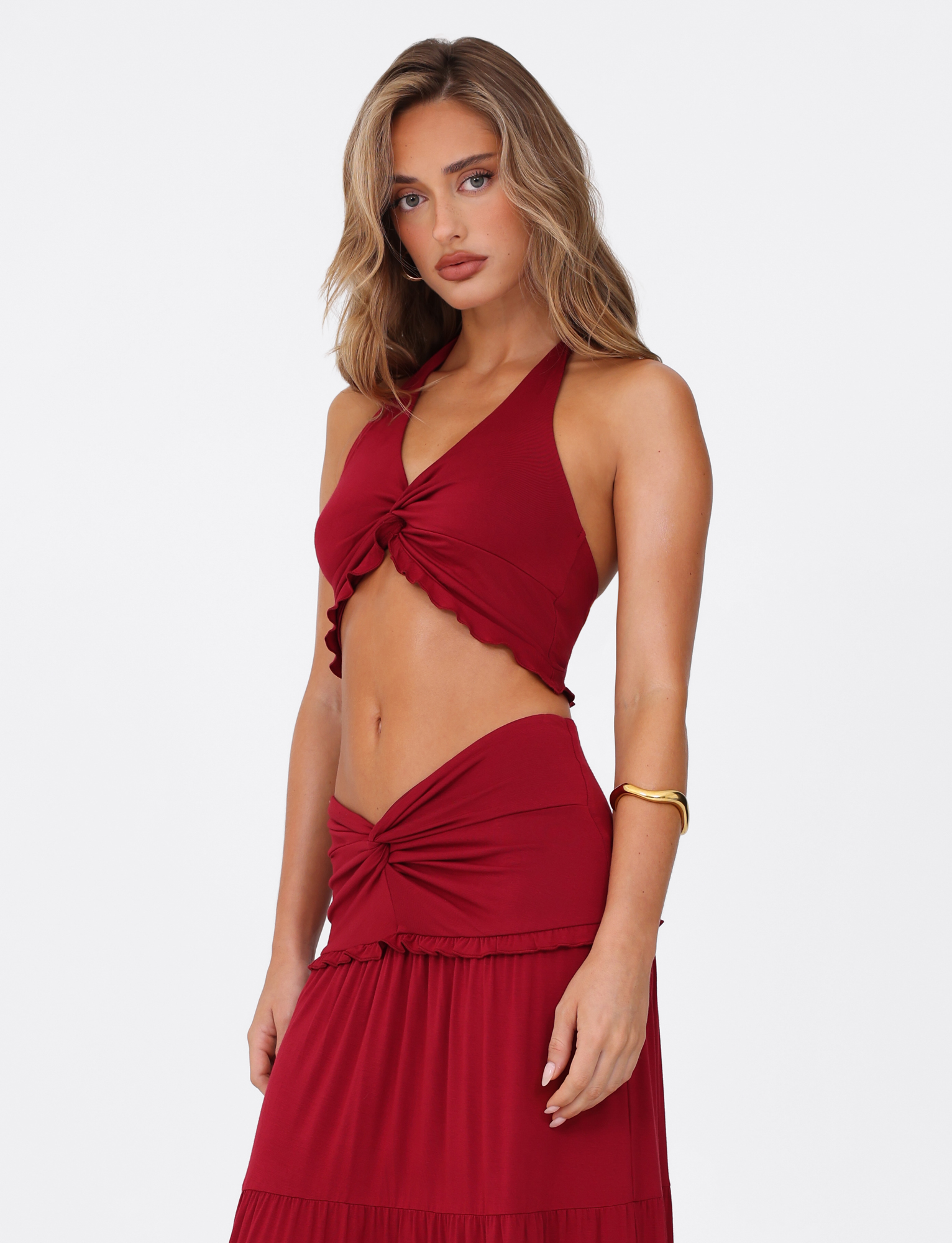 A woman with wavy light brown hair wears the TIGERMIST KATYA HALTER TOP in red, featuring a sleeveless, halter neck and twist detail. She stands against a white background, looking at the camera with a neutral expression.