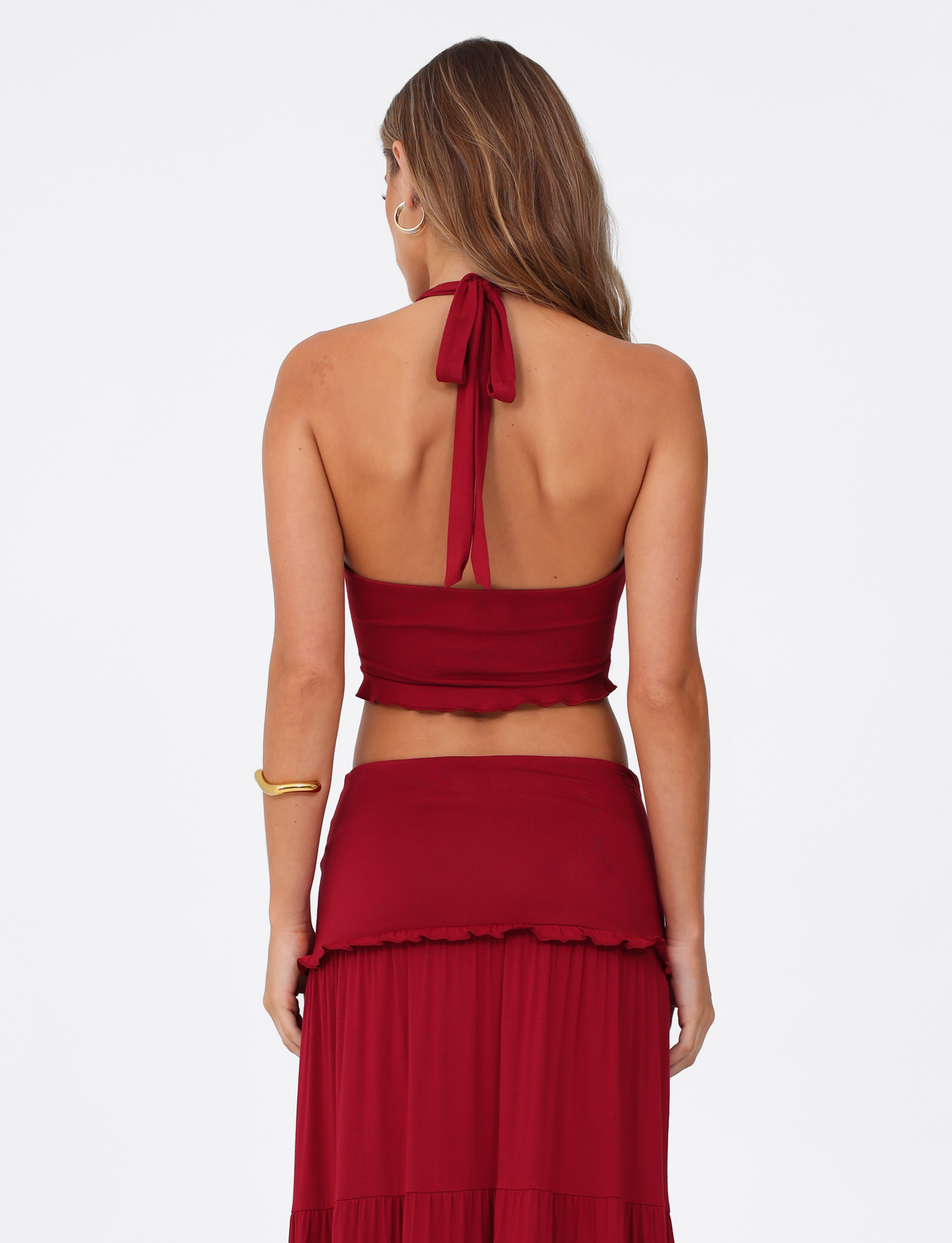 A woman with long brown hair is shown from the back wearing the TIGERMIST KATYA HALTER TOP in red and a matching skirt, accessorized with a gold bracelet on her upper right arm. The background is plain white.
