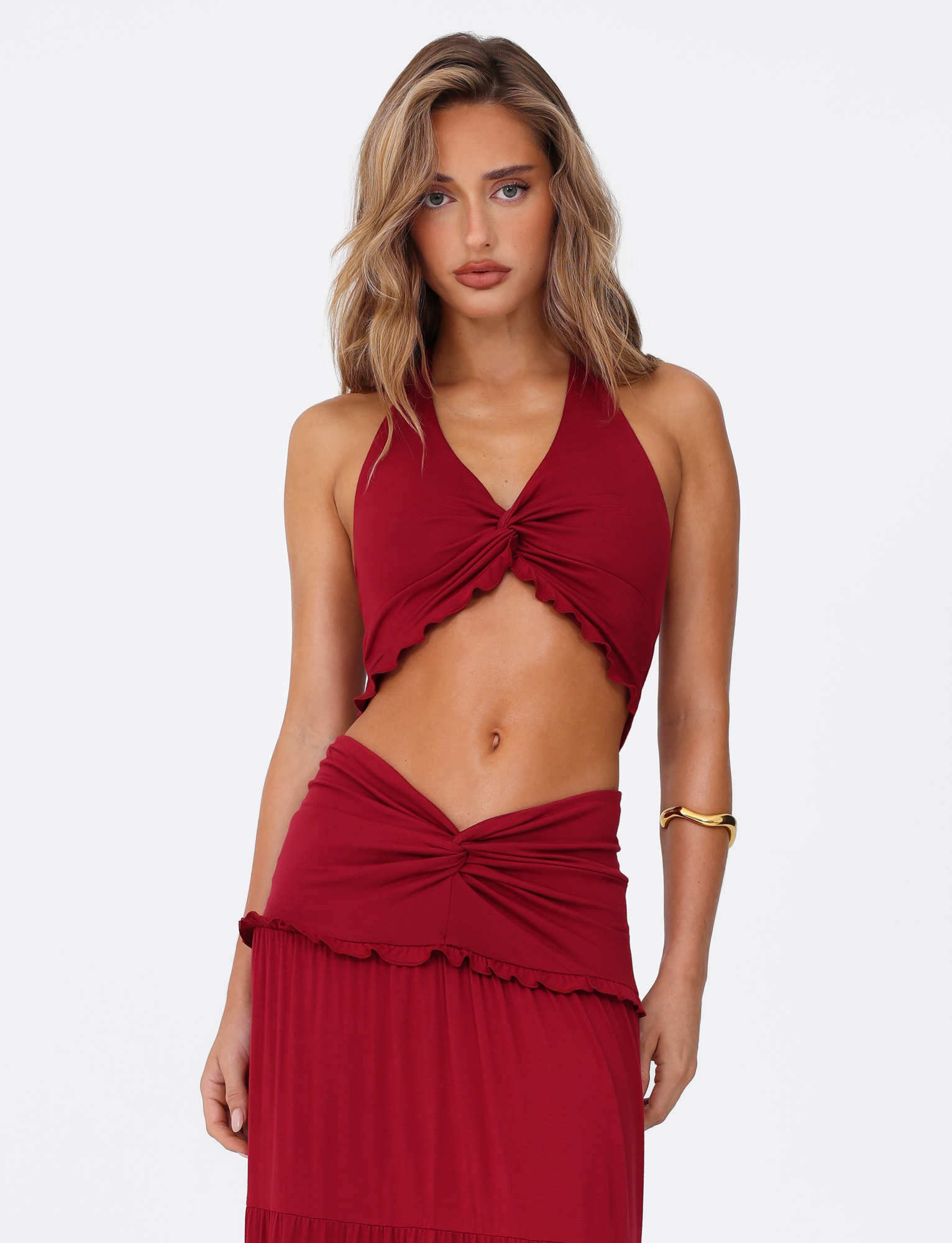 A woman with wavy blonde hair wears the TIGERMIST KATYA HALTER TOP in red, a sleeveless crop with a halter neck, paired with a matching skirt featuring ruched and knotted front details. She completes the look with a gold upper arm bracelet.