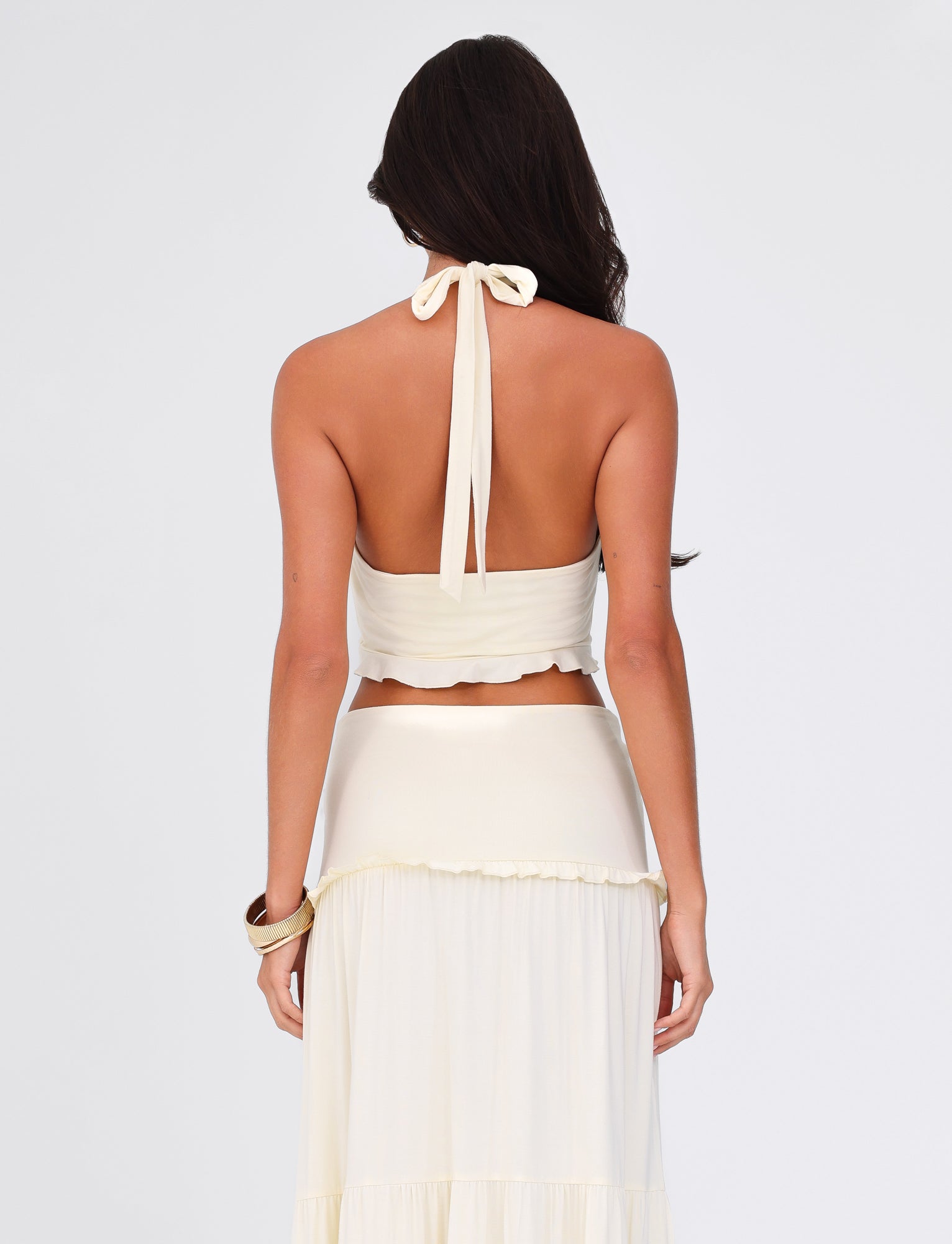 A woman with long dark hair wears the TIGERMIST KATYA HALTER TOP in cream, showcasing the twist detail of her buttery jersey ensemble. She pairs it with a light, flowing skirt and holds her bracelet-adorned arm against a plain background.