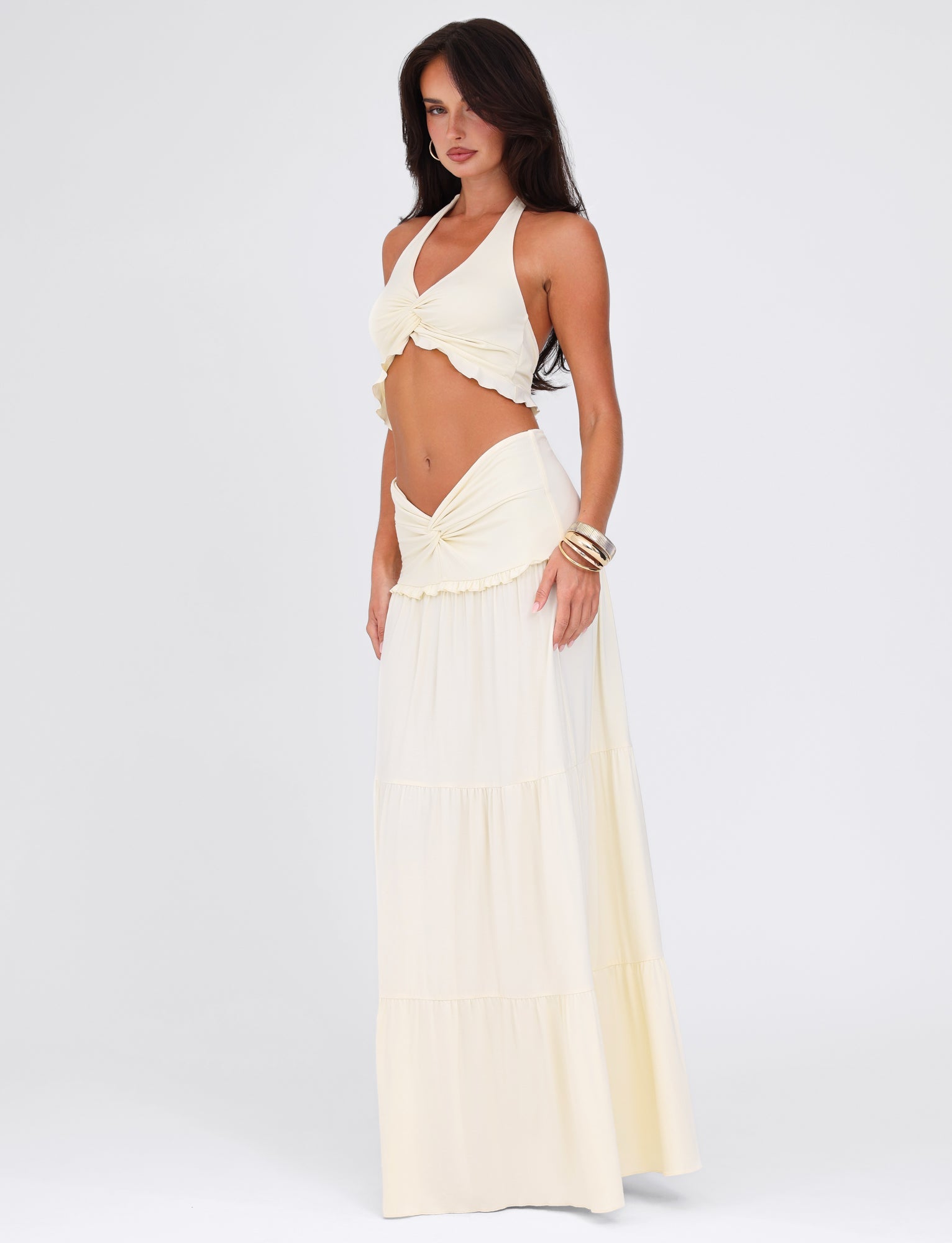 A woman poses in the TIGERMIST KATYA MAXI SKIRT - WHITE : CREAM, complemented by a cream halter top. She stands against a white backdrop, adorned with a wrist bangle and her long hair cascading down.