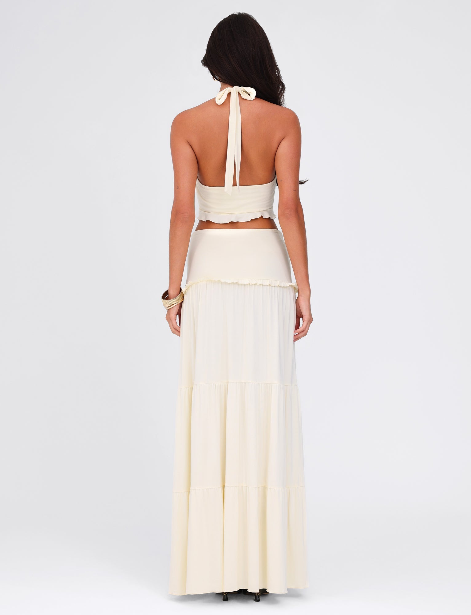 A woman models the TIGERMIST KATYA MAXI SKIRT in cream, paired with a backless halter top. She stands with her back to the camera, highlighting the skirt's subtle twist detail and high stretch fabric against a plain white background.