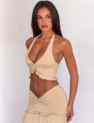 A woman with long dark hair wears the TIGERMIST KATYA TOP in tan, featuring a halter neck and twist detail. She stands against a plain light background, looking confidently at the camera.