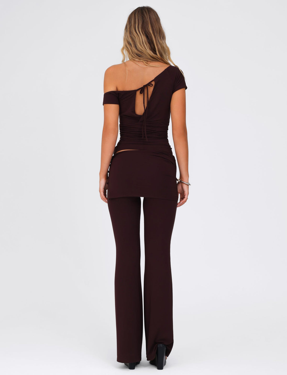 A woman with long, wavy hair is wearing the TIGERMIST dark-brown, off-the-shoulder top featuring a back tie detail and the LEILANI PANT - BROWN : CHOCOLATE flared leg pants made of high stretch fabric. She stands on a plain white background, facing away from the camera.