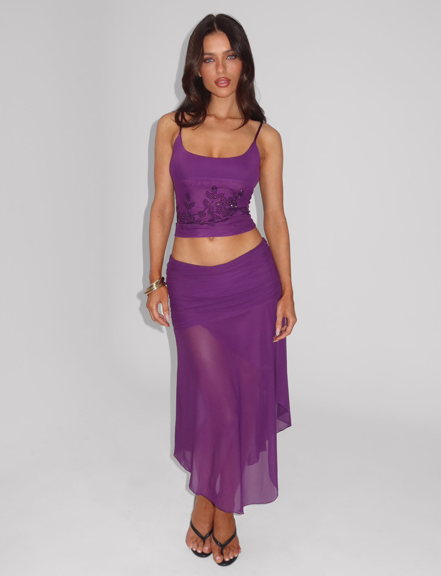 A woman with long dark hair wears the TIGERMIST MARIANA SKIRT - PURPLE and a fitted purple tank top with floral embroidery. She stands against a plain white background, styling the look with black heeled sandals and a gold bracelet.