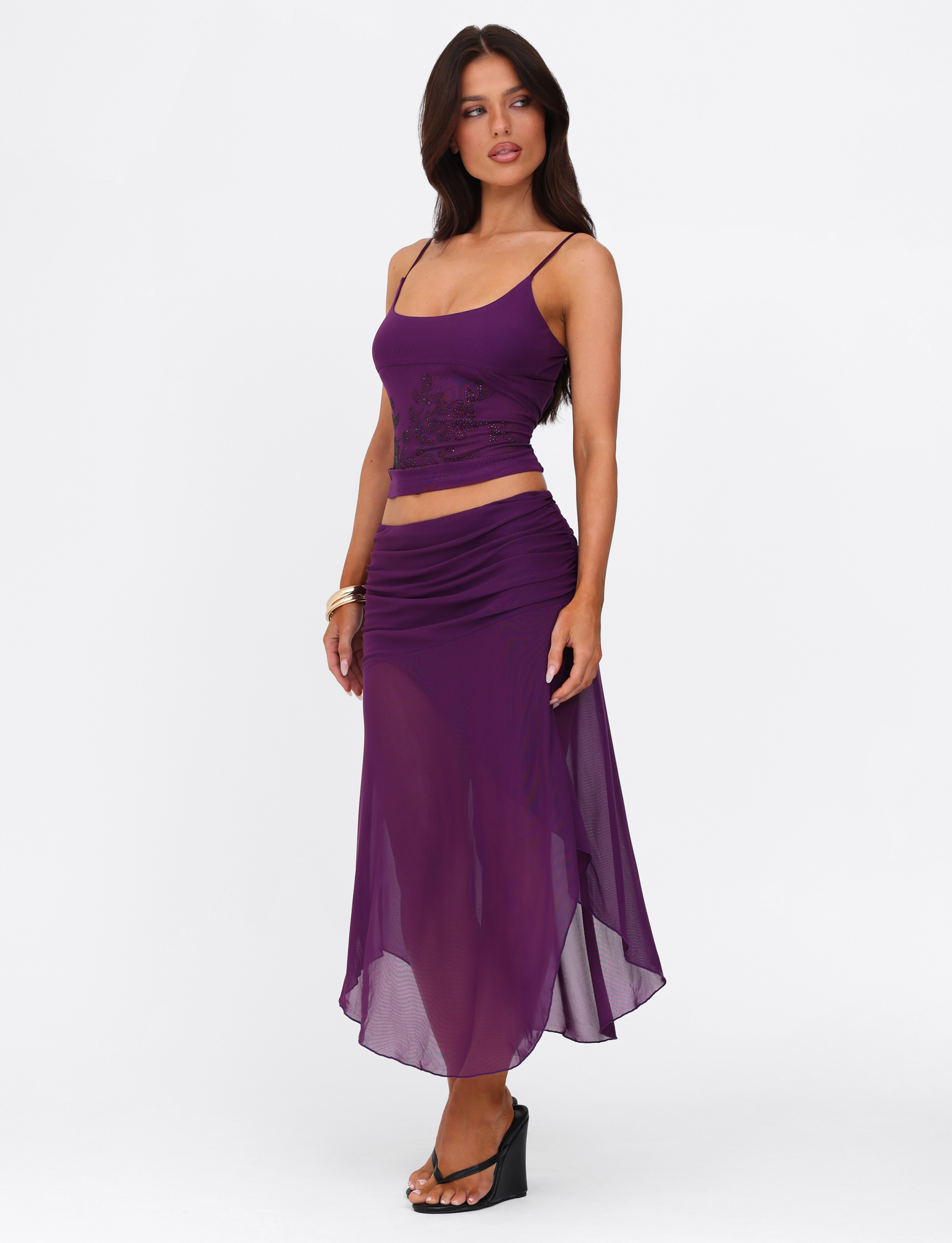 A woman stands against a white background, looking slightly to the side, wearing a sleeveless purple top and the TIGERMIST MARIANA SKIRT - PURPLE, a sheer asymmetric wrap midi skirt, paired with sandals.