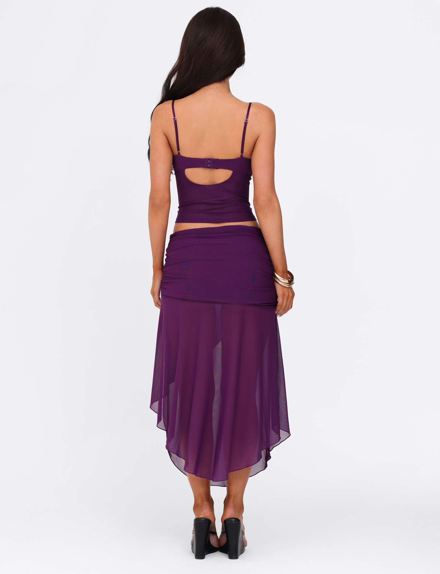 A woman with long brown hair faces away, wearing a fitted purple sleeveless top and the TIGERMIST MARIANA SKIRT - PURPLE, a sheer asymmetric wrap midi skirt, paired with black heels and gold bracelets against a white background.