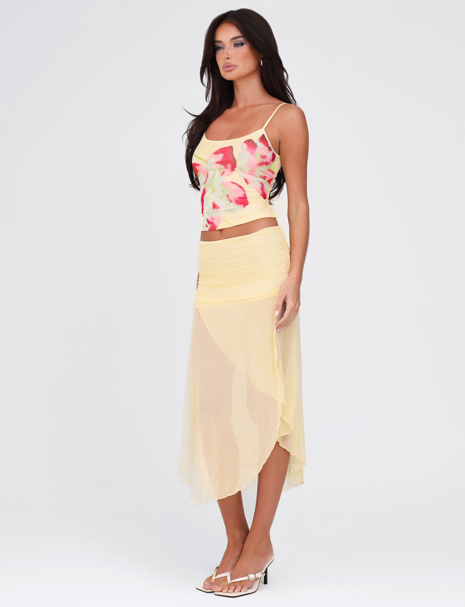 A woman with long brown hair and a neutral expression wears the TIGERMIST MARIANA SKIRT in pale yellow against a plain white background, paired with a colorful floral tank top and white heeled sandals.