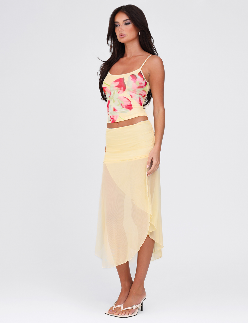A woman with long brown hair and a neutral expression wears the TIGERMIST MARIANA SKIRT in pale yellow against a plain white background, paired with a colorful floral tank top and white heeled sandals.