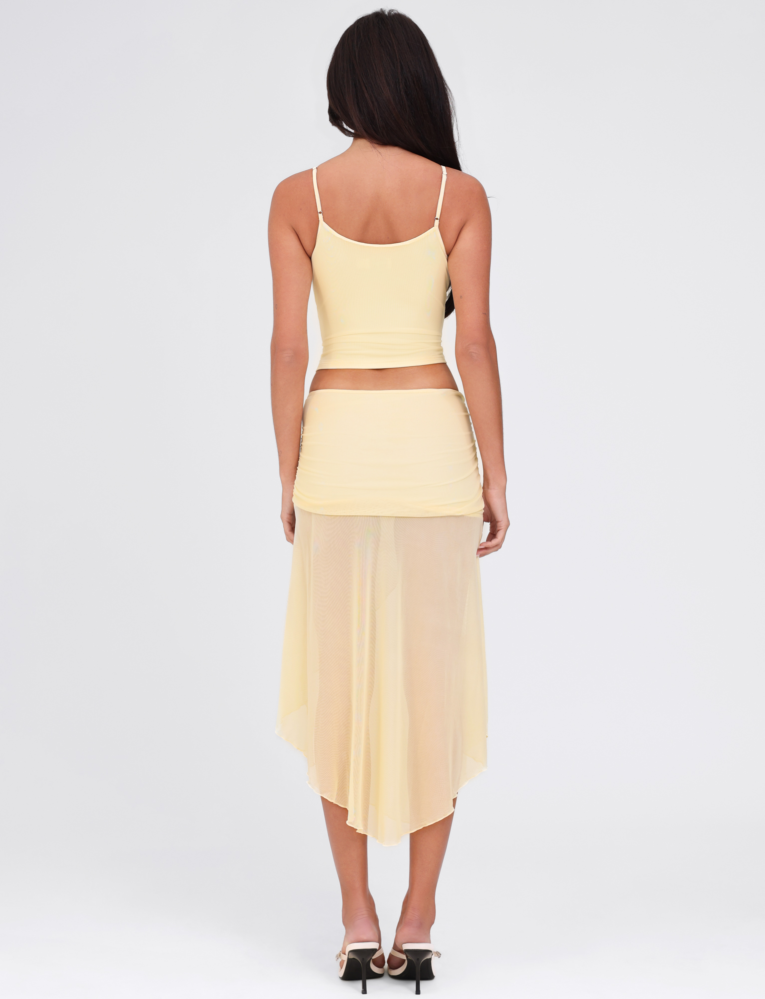 A woman with long dark hair faces away from the camera, wearing the TIGERMIST MARIANA SKIRT in pale yellow and a matching light yellow spaghetti strap top, set against a plain white background.