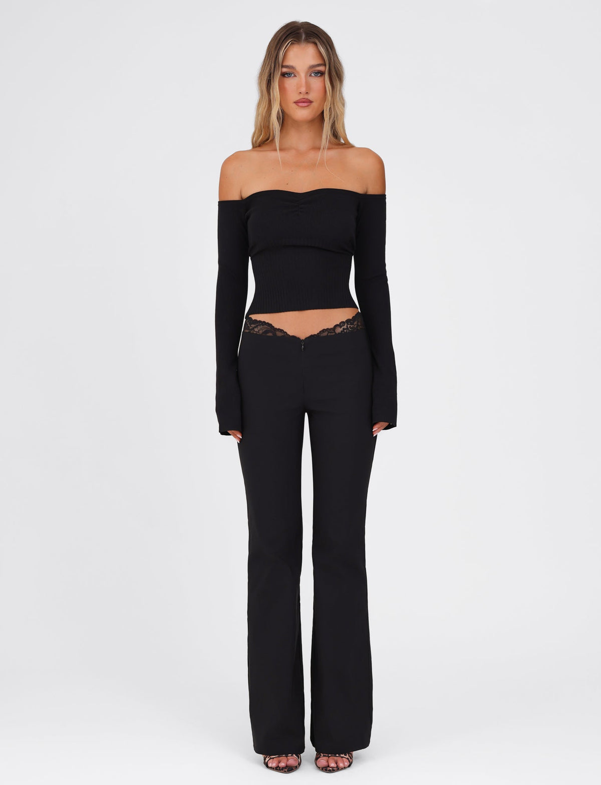 A woman stands against a plain background, showcasing the LILYANA LONG SLEEVE TOP in black from TIGERMIST. This elegant off-the-shoulder, fully fashioned knit is fitted through the body and sleeves, paired with black pants that feature lace detailing. Her hair cascades loosely, and she completes the ensemble with open-toe shoes.