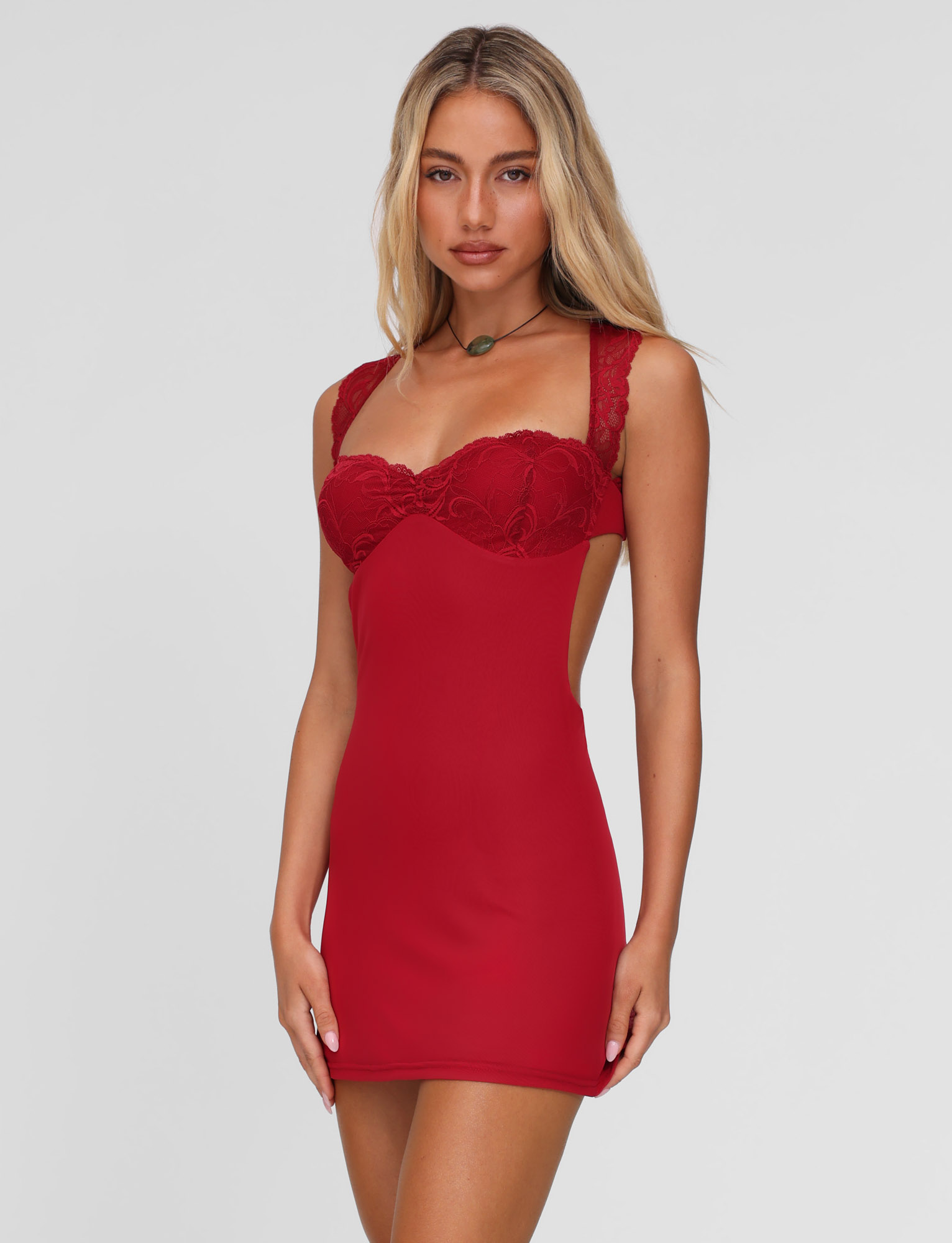 A woman with long blonde hair wears the TIGERMIST NYOMI MINI DRESS - RED, a fitted, sleeveless red mini dress with a lace bust panel and a necklace, standing against a plain light background.