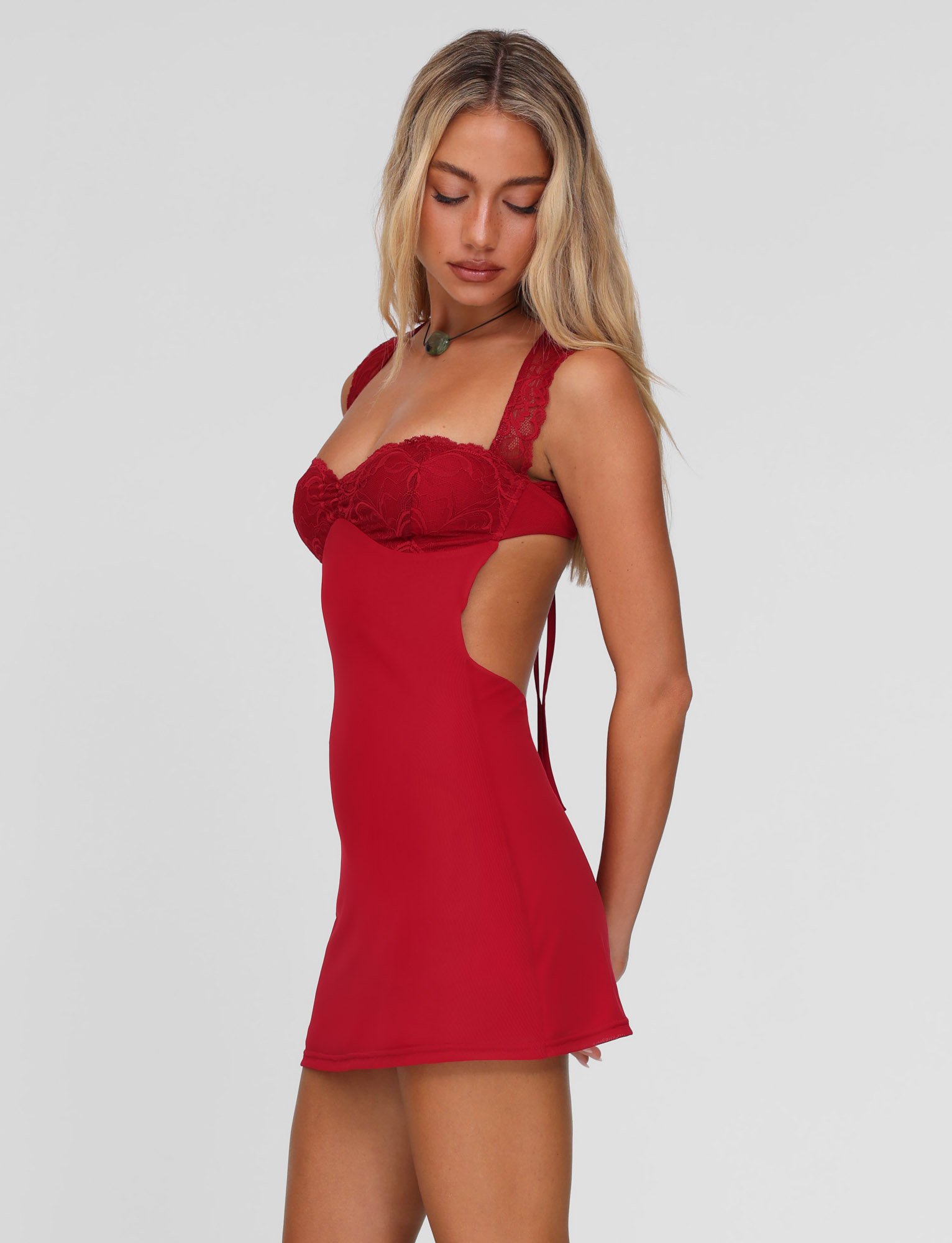 A woman with long blonde hair wears the TIGERMIST NYOMI MINI DRESS in red, featuring a lace bust panel and open sides, as she looks down neutrally against a plain light background.