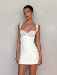 A woman stands against a light beige wall wearing the TIGERMIST NYOMI MINI DRESS in white and cream. Her dark hair is pulled back as she accessorizes with small hoop earrings and a bracelet, gazing slightly to her left.