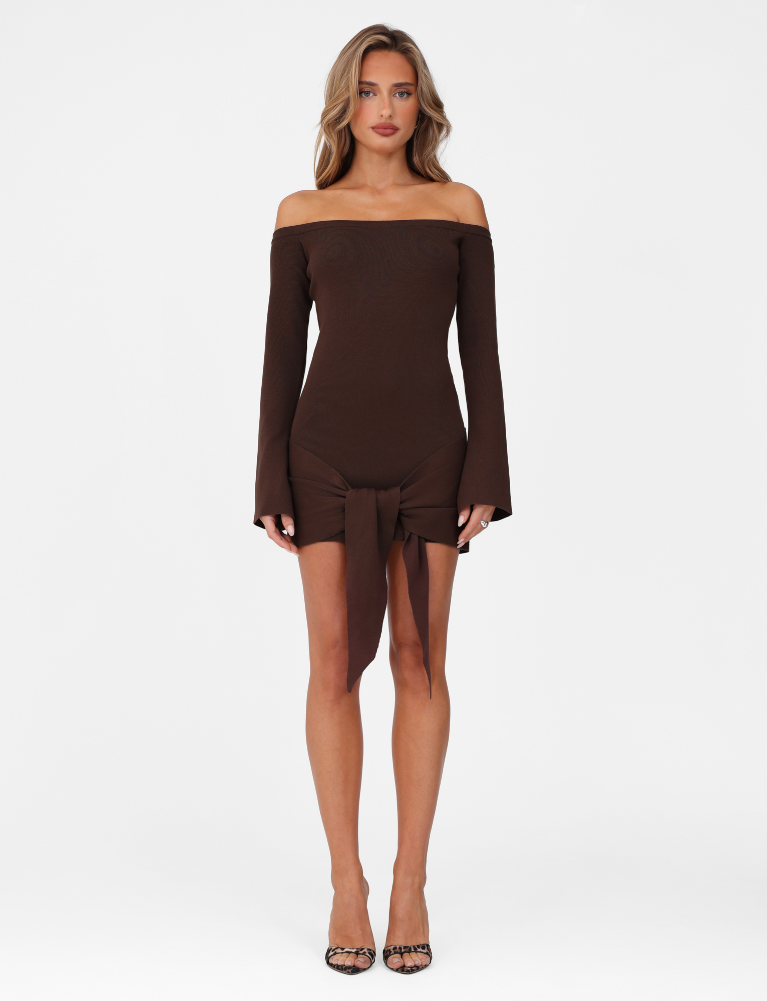 A woman with wavy hair and a neutral expression stands against a white background, wearing the TIGERMIST CYPRESS MINI DRESS in chocolate brown. The dress features an off-the-shoulder neckline, long sleeves, and a front tie detail, paired with open-toe heels.