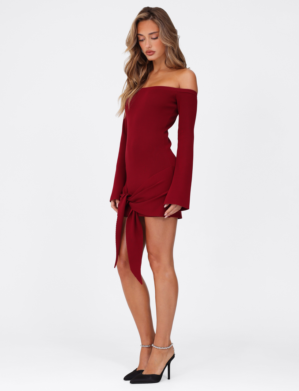 A woman stands in profile wearing the TIGERMIST CYPRESS MINI DRESS - RED : WINE, featuring an elasticated off-the-shoulder neckline, long sleeves, and a front knot detail, paired with black pointed-toe heels against a white background.