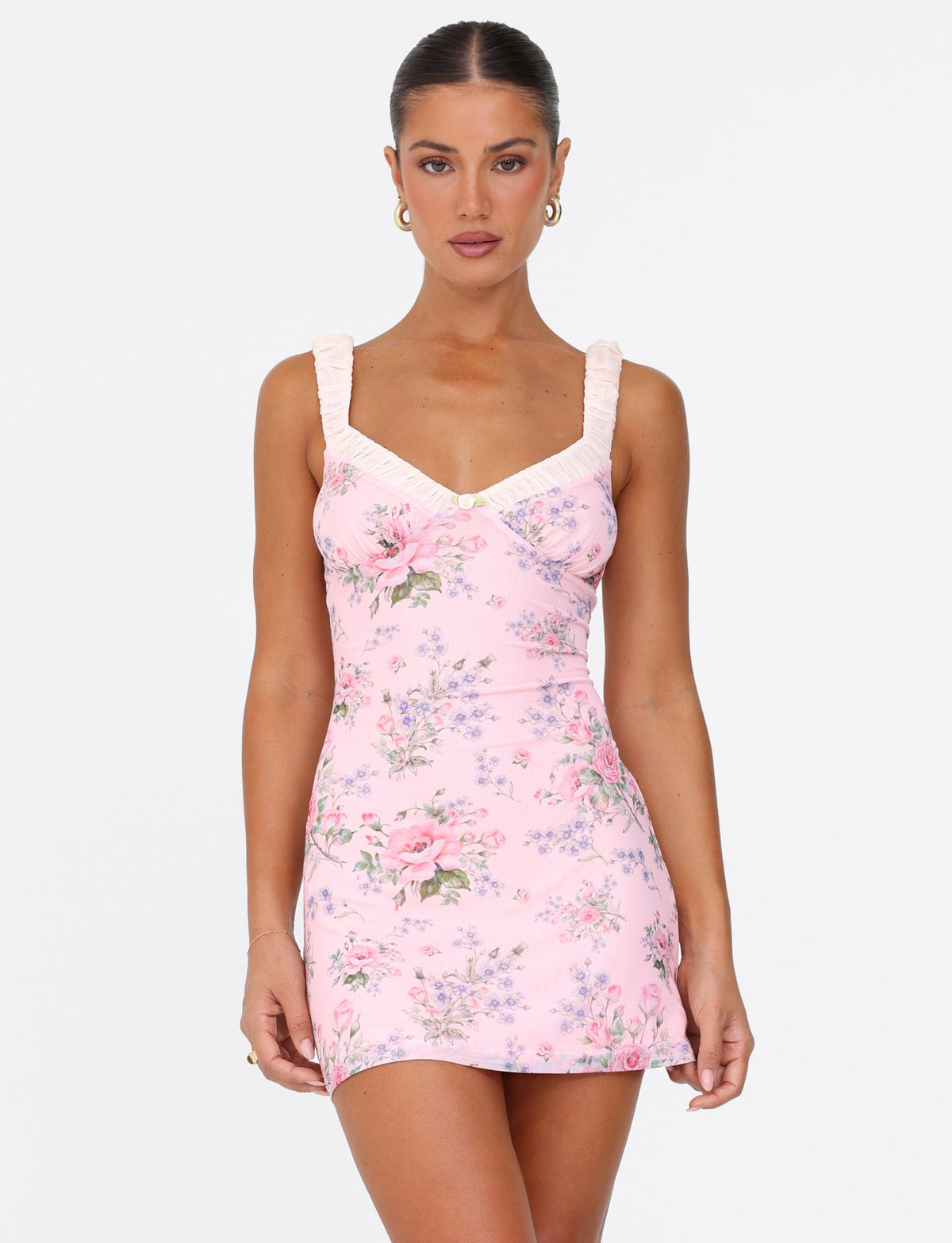A woman wearing the AMARAN MINI DRESS by TIGERMIST in a pink wild flower print with bustier styling and ruffled straps poses against a plain white background. Her hair is tied back, and she accessorizes with hoop earrings while she gazes directly at the camera with a neutral expression.