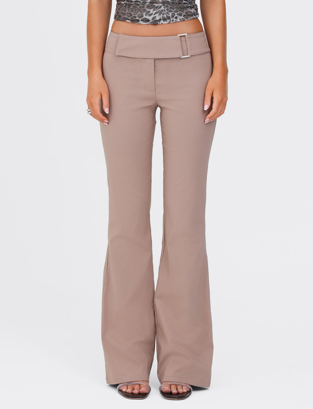 A person stands against a white background wearing the VESPER PANT - BROWN : TAN by TIGERMIST. The taupe-colored flare pants feature a wide waistband and a rectangular belt buckle. The solid tan pants have a full-length silhouette that complements their patterned cropped top. They are wearing sandals, with their hands resting by their sides.