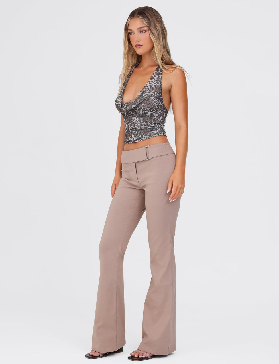 A woman with long, wavy hair is standing against a plain white background. She is wearing a sleeveless, low-cut, patterned top and the VESPER PANT in TAN from TIGERMIST. The pants feature a high-waisted belt and flared legs. She looks off to the side and is wearing open-toed black shoes.