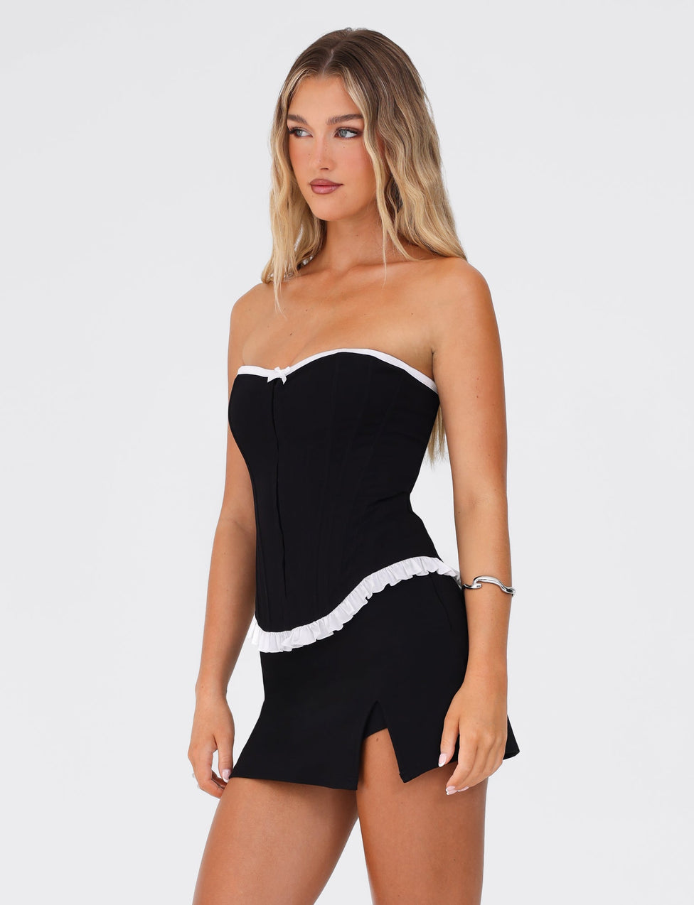 A woman with long blonde hair is wearing the TIGERMIST MILLIE CORSET 2.0 - BLACK, a black strapless corset top with white trim and a matching black mini skirt featuring a side slit and bodycon fit. She is standing against a plain white background, looking off to the side, and has a silver bracelet on her right wrist.