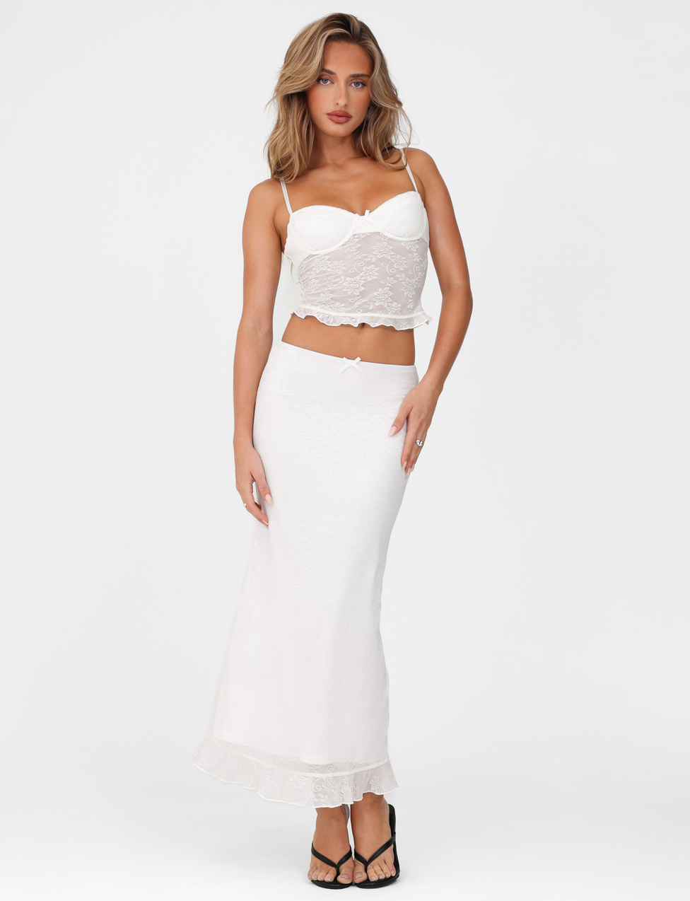 A woman with wavy light brown hair wears the TIGERMIST CHARLEIGH TOP - WHITE LACE, a white lace camisole with thin straps, paired with a long matching skirt and black sandals, standing against a plain white background.