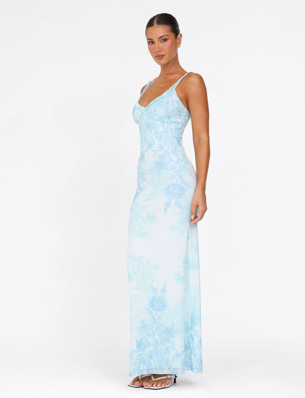 A woman wearing the TIGERMIST GUILIANNA FRILL MAXI DRESS in a light blue floral tapestry pattern stands against a plain white background. The dress features frill shoulder straps, which she complements with strappy white heels and her hair styled in a tied-back manner. Her pose is relaxed, facing slightly to the side.