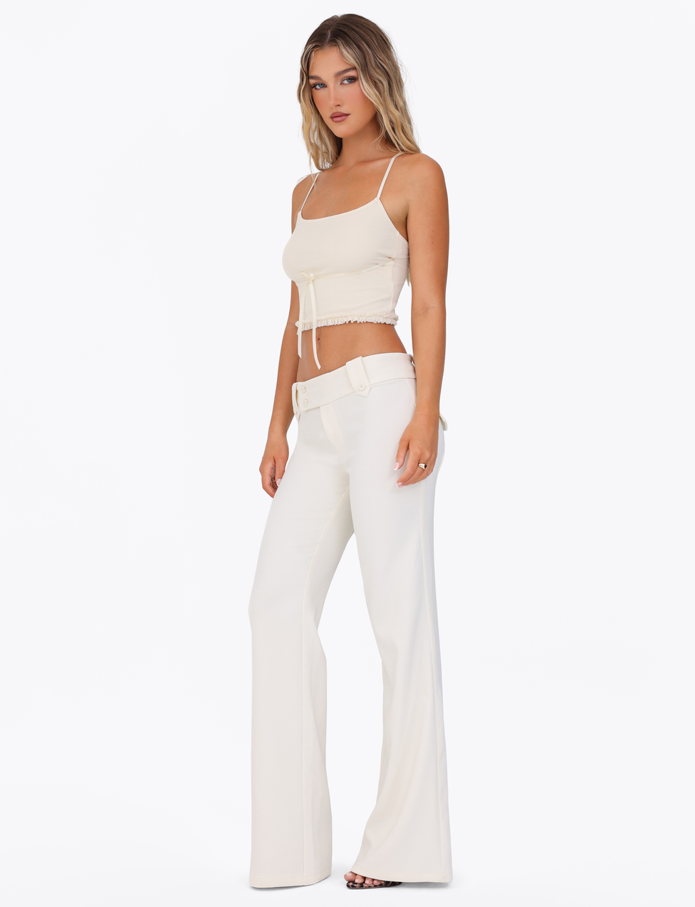 A woman faces slightly sideways against a plain white background, wearing TIGERMIST’s KITTIE PANT in white/cream paired with a light beige cropped tank top with thin straps and tie detail, and open-toe heels.