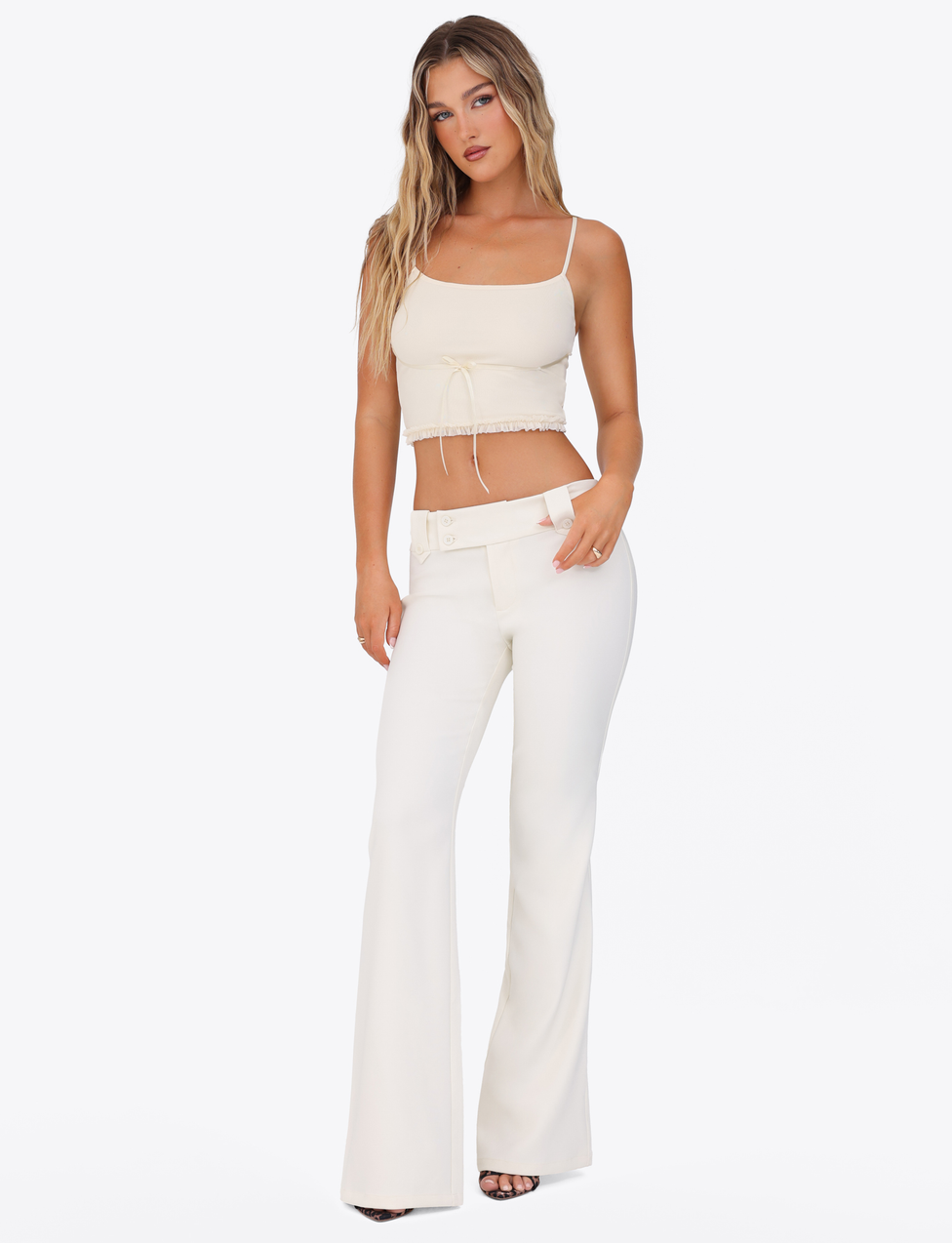 A woman with long, wavy hair poses against a white background wearing TIGERMIST’s KITTIE PANT - WHITE : CREAM, a fitted, high-waisted flared pant paired with a matching cream spaghetti strap crop top.