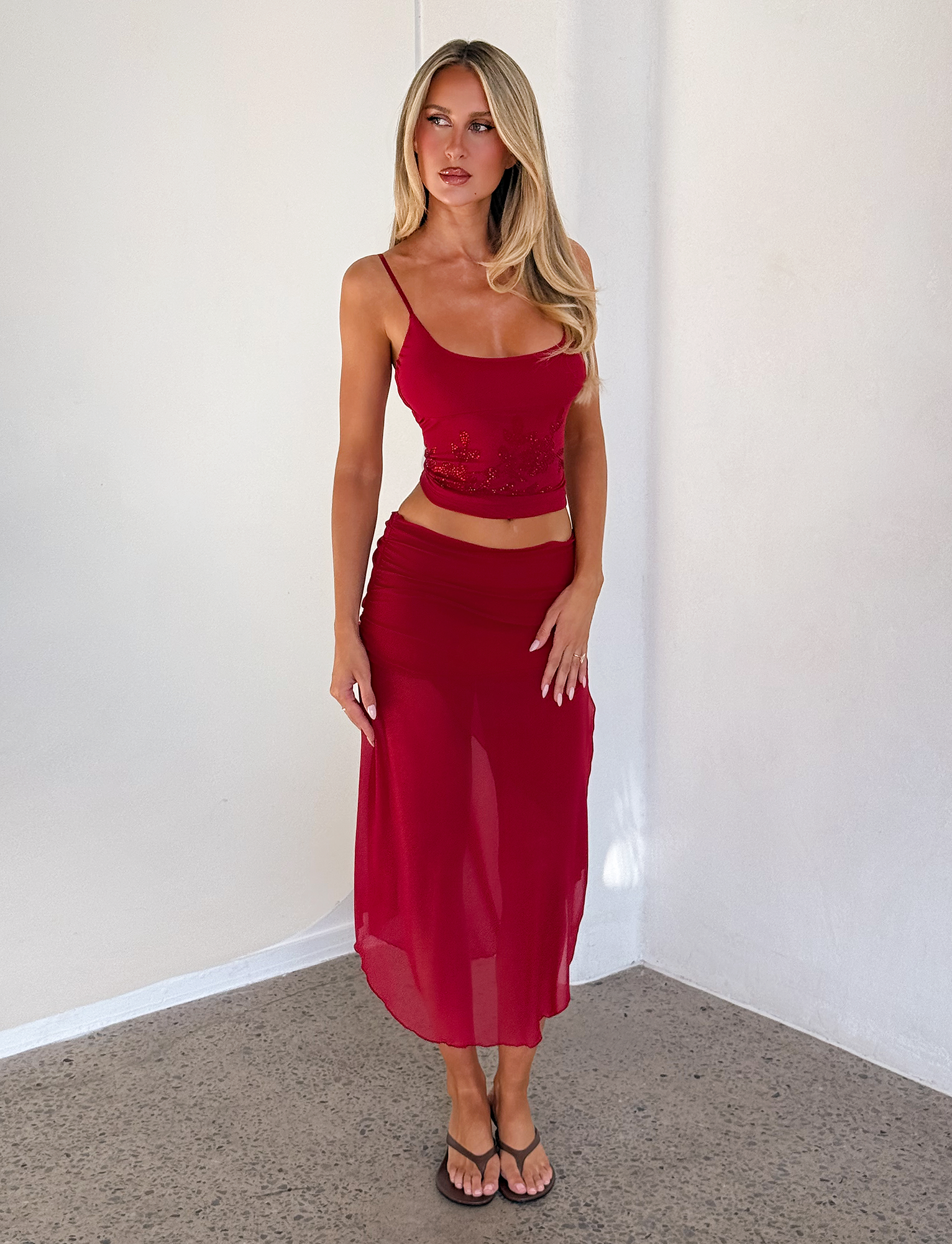A woman with long blonde hair wears the TIGERMIST OCTAVIA TOP in red and a matching sheer skirt, standing indoors by a light-colored wall and floor, paired with black sandals.