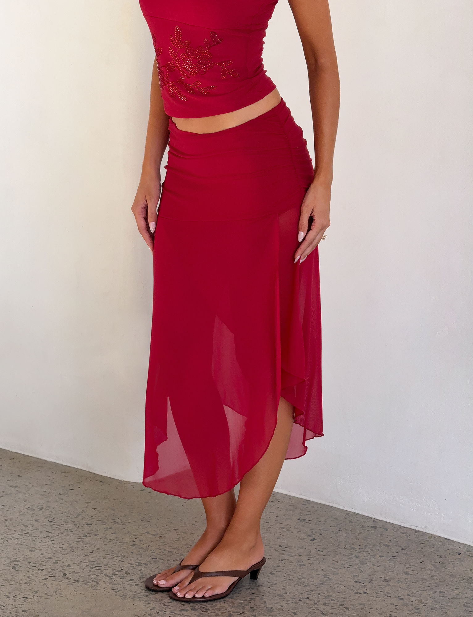 A person wears a sleeveless red top and the TIGERMIST MARIANA SKIRT - RED, featuring a ruched waistband and asymmetric wrap design, paired with black sandals. They stand against a plain light-colored wall. The head is not shown in the image.