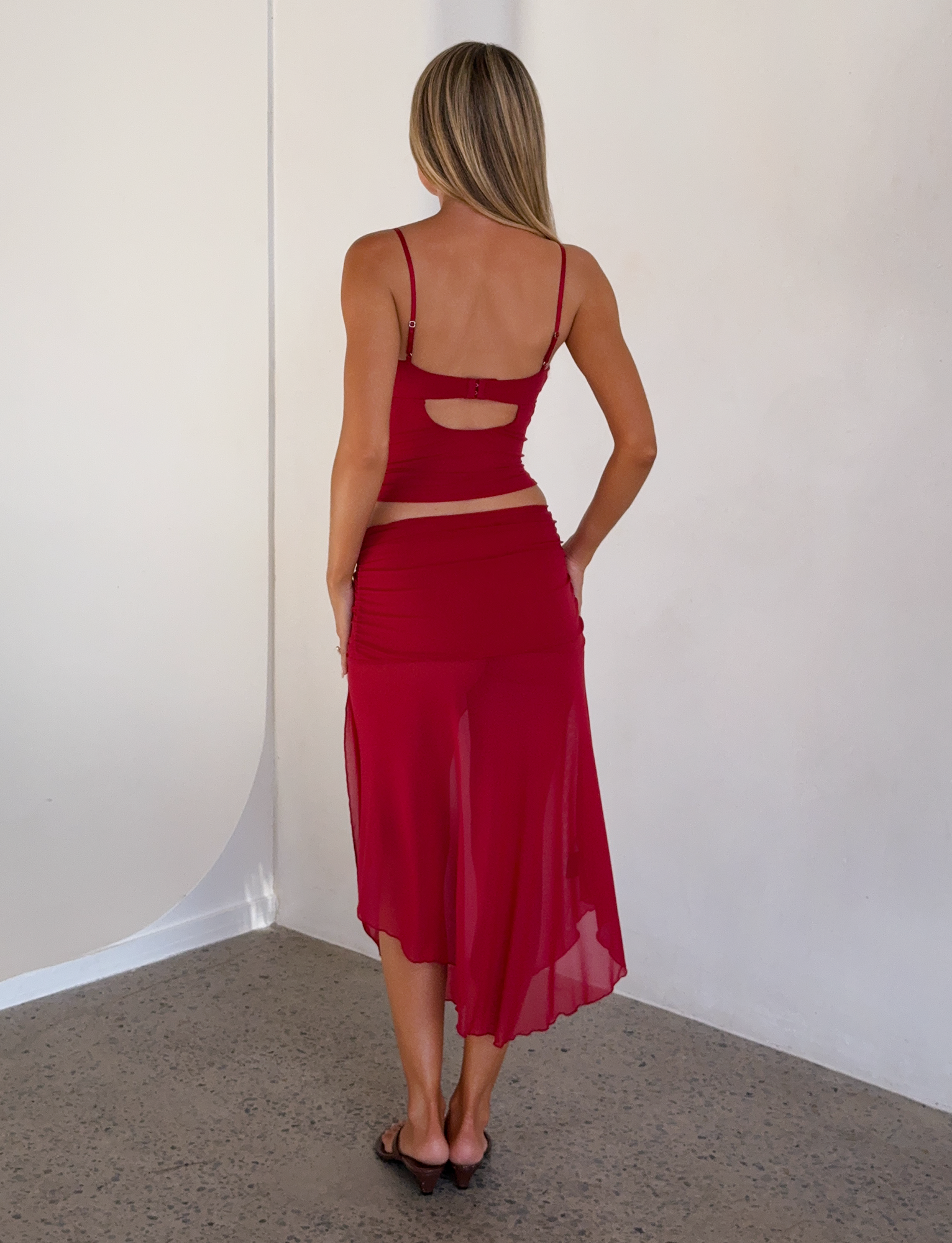 A woman with long blonde hair faces away, wearing a red sleeveless crop top, sandals, and the TIGERMIST MARIANA SKIRT - RED—a sheer red skirt with a ruched waistband—in a minimalist room with white walls and a speckled floor.