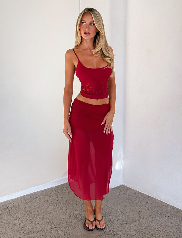 A woman with long blonde hair wears the Tiger Mist USA CL - MARIANA SKIRT, styled with a sleeveless red crop top and black sandals, standing against a plain white wall on a concrete floor.