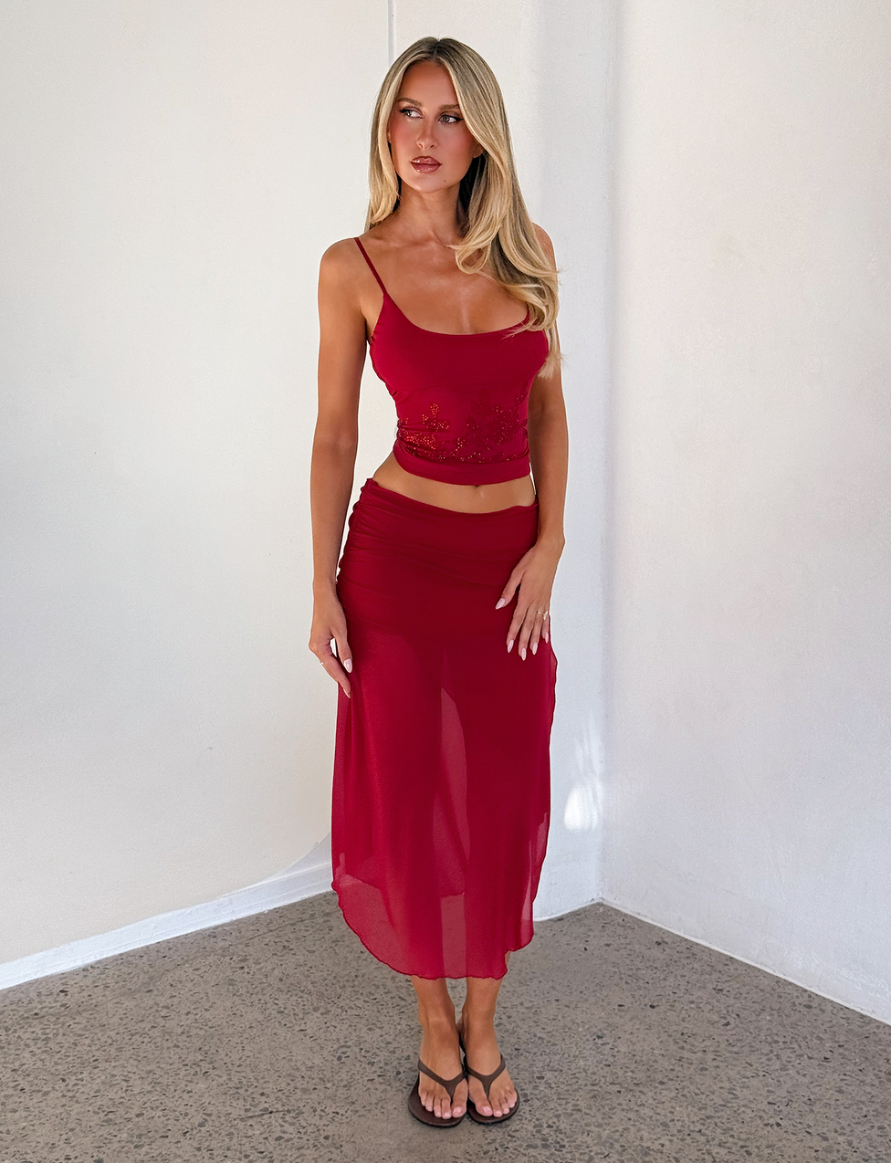 A woman with long blonde hair wears the Tiger Mist USA CL - MARIANA SKIRT, styled with a sleeveless red crop top and black sandals, standing against a plain white wall on a concrete floor.