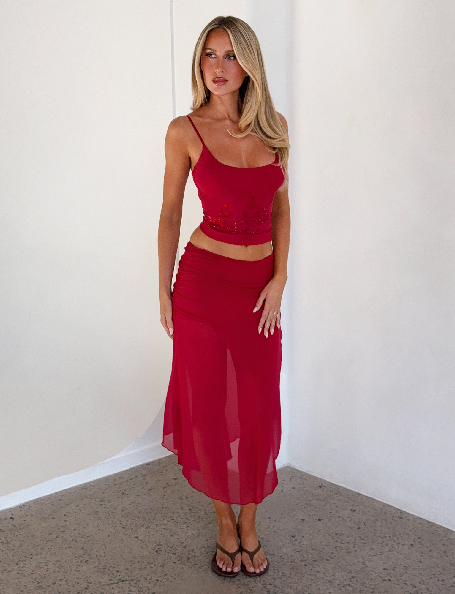 A woman with long blonde hair wears the TIGERMIST OCTAVIA TOP in red and a matching sheer red skirt, paired with black sandals. She stands indoors against a white wall, gazing slightly to the side.