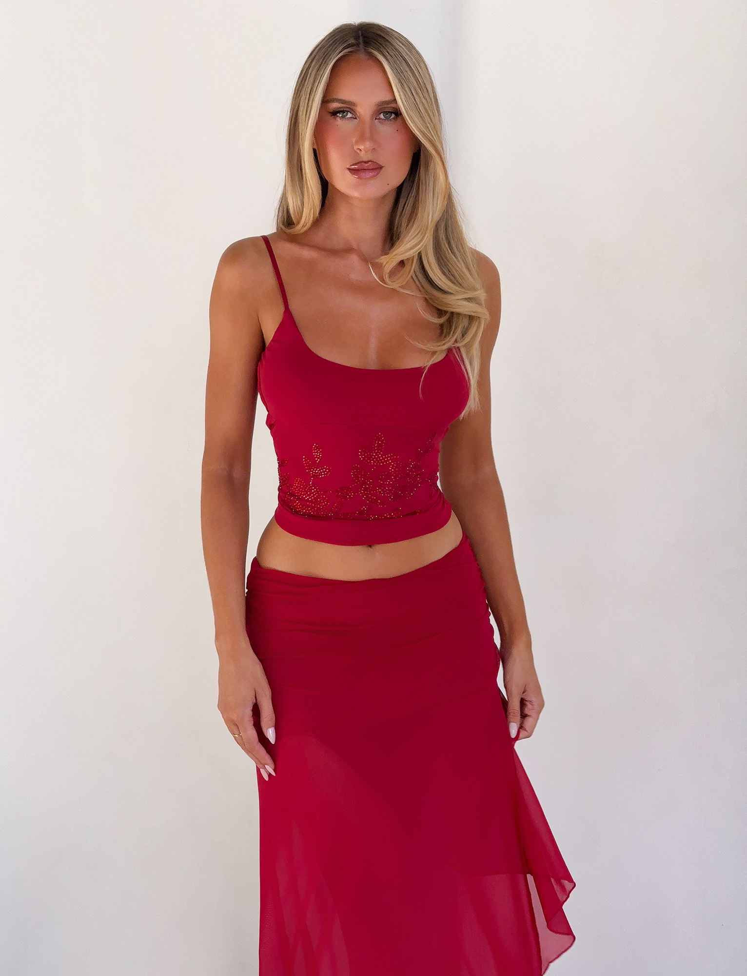 A woman with long blonde hair stands against a white wall, wearing a red sleeveless top and the TIGERMIST MARIANA SKIRT - RED, featuring an asymmetric wrap design. She looks directly at the camera with a neutral expression.