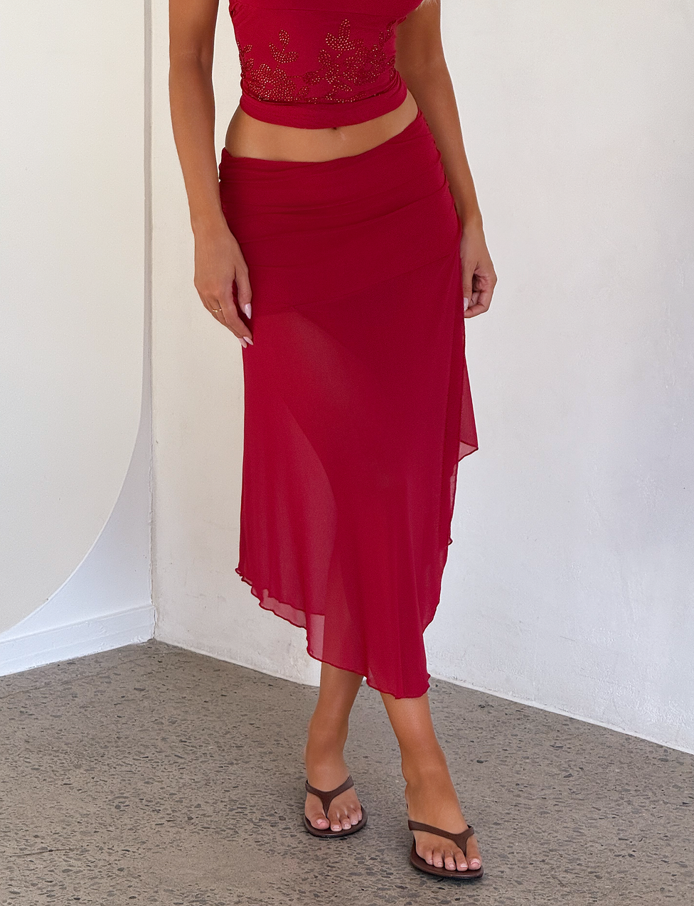 A woman stands indoors on a gray floor wearing black sandals, a sleeveless red crop top, and the TIGERMIST MARIANA SKIRT - RED with an asymmetric wrap design. Her face is not visible, and one hand is by her side.