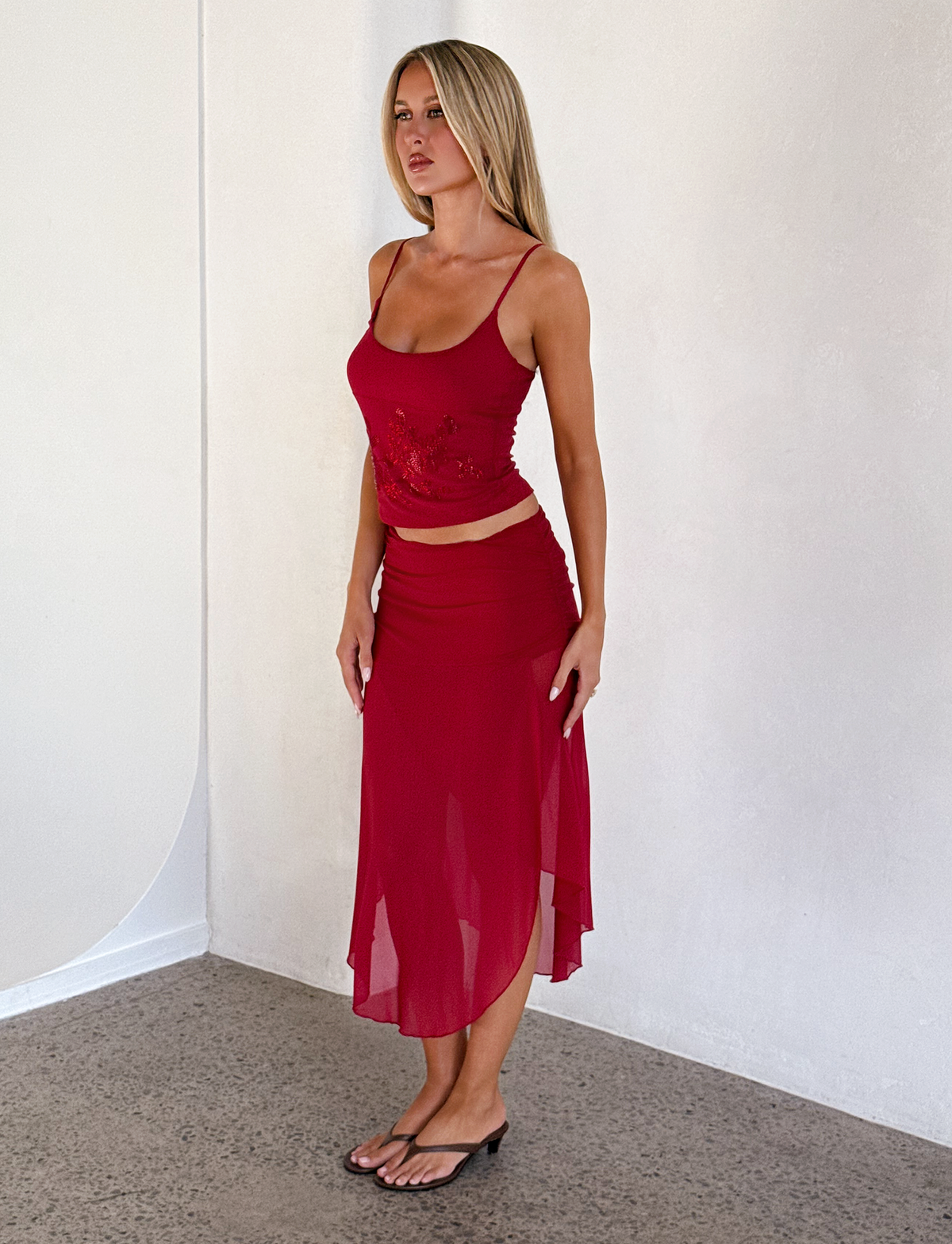 A woman with long blonde hair stands indoors by a light wall, wearing a sleeveless red top and the TIGERMIST MARIANA SKIRT - RED, a flowy asymmetric wrap style with ruched waistband, paired with flip-flops. She faces slightly left with a neutral expression.