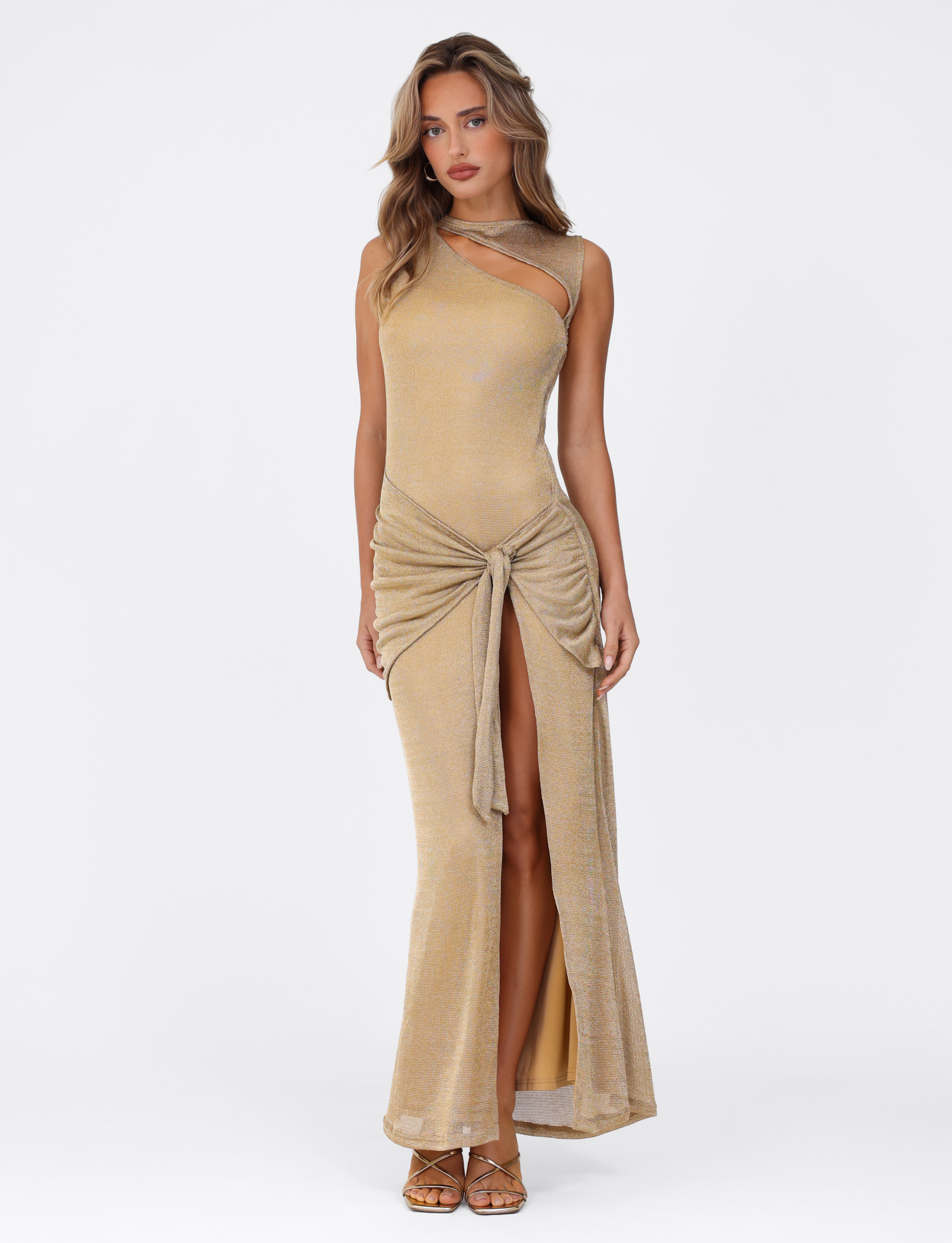 A woman poses against a white background in the TIGERMIST AZELMA MAXI DRESS - GOLD, a metallic knit dress with a maxi silhouette, high slit, and knotted waist detail. She pairs it with gold heels and wears her hair wavy.