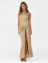 A woman poses against a white background in the TIGERMIST AZELMA MAXI DRESS - GOLD, a metallic knit dress with a maxi silhouette, high slit, and knotted waist detail. She pairs it with gold heels and wears her hair wavy.