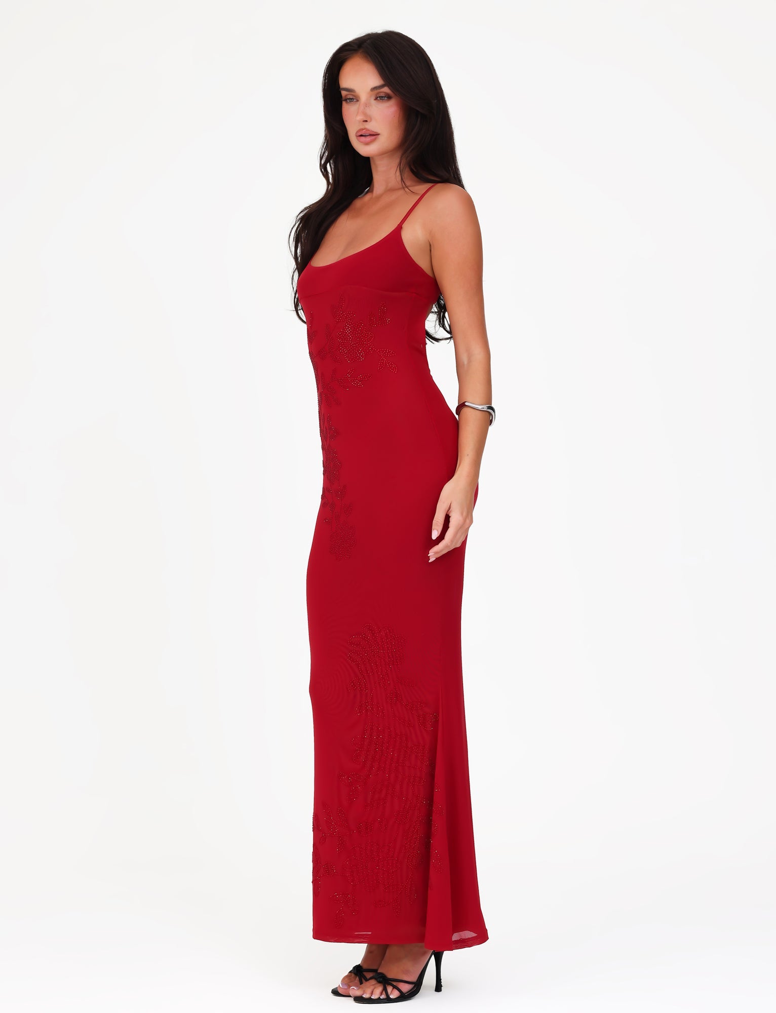 A woman in a striking Cherry Red Octavia Maxi Dress by Tigermist stands gracefully against a plain white background. She pairs the dress with black high-heeled sandals and a delicate bracelet, while her long hair cascades elegantly down her back.