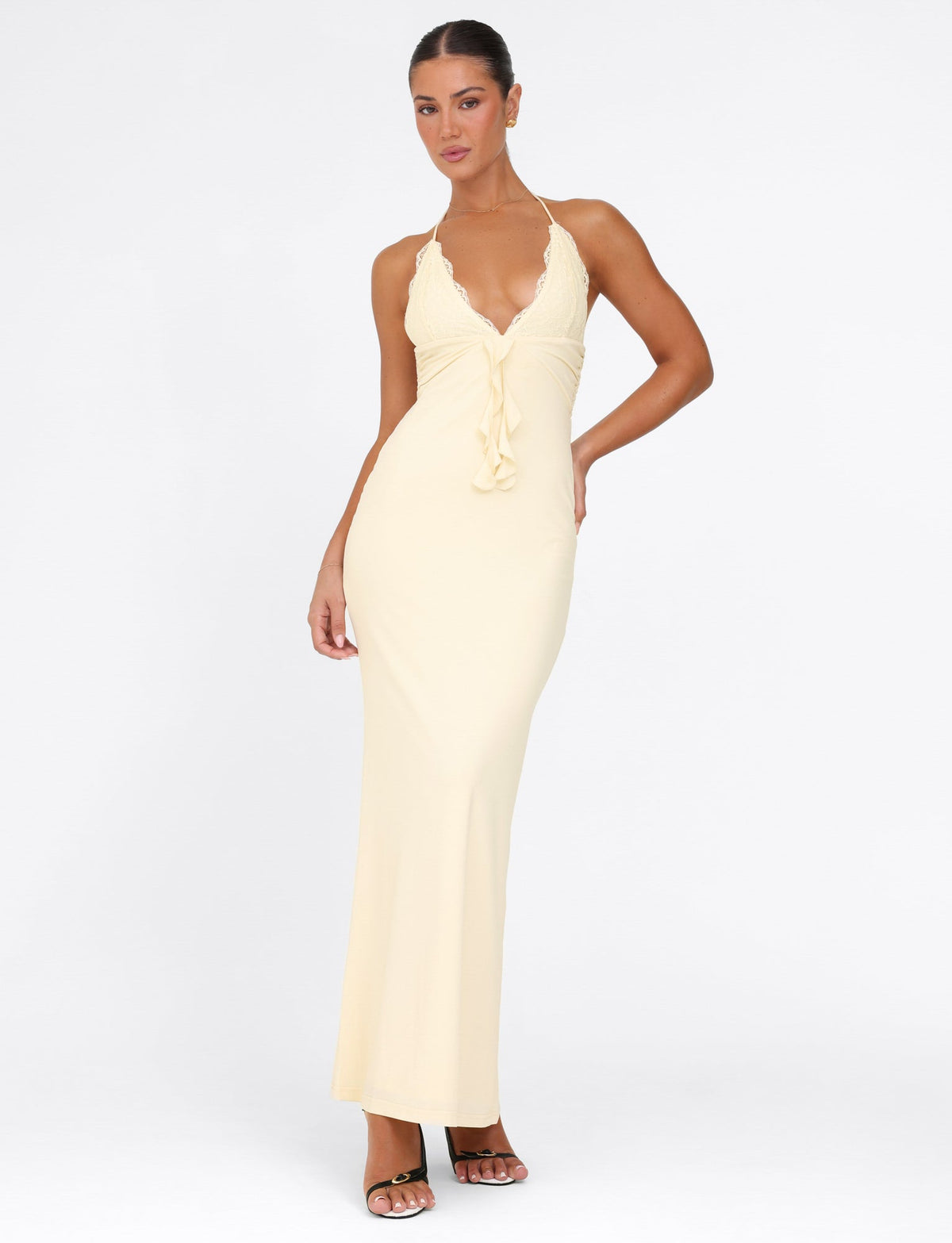 A woman poses confidently in the chic Deena Maxi Dress by Tigermist, an elegant lemon-yellow ensemble featuring a plunging neckline and halter top. Lace bust cups enhance its elegance as she stands in black high-heeled sandals against a plain white background.