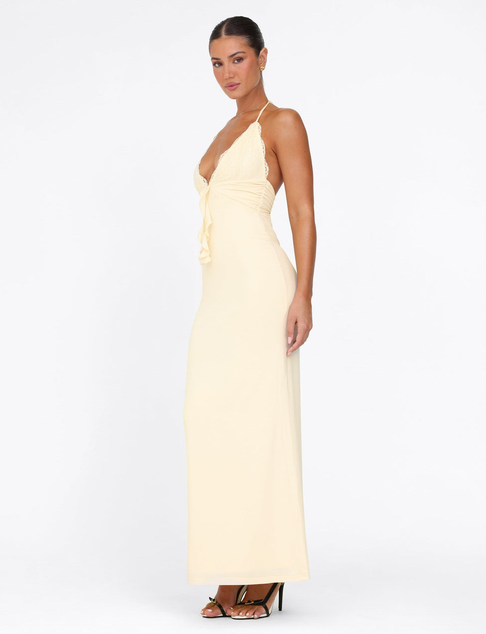 A person poses sideways in the long, lemon yellow Deena Maxi Dress from TIGERMIST, featuring lace bust cups. Their hair is pulled back, and they are wearing black high-heeled sandals against a plain white background.