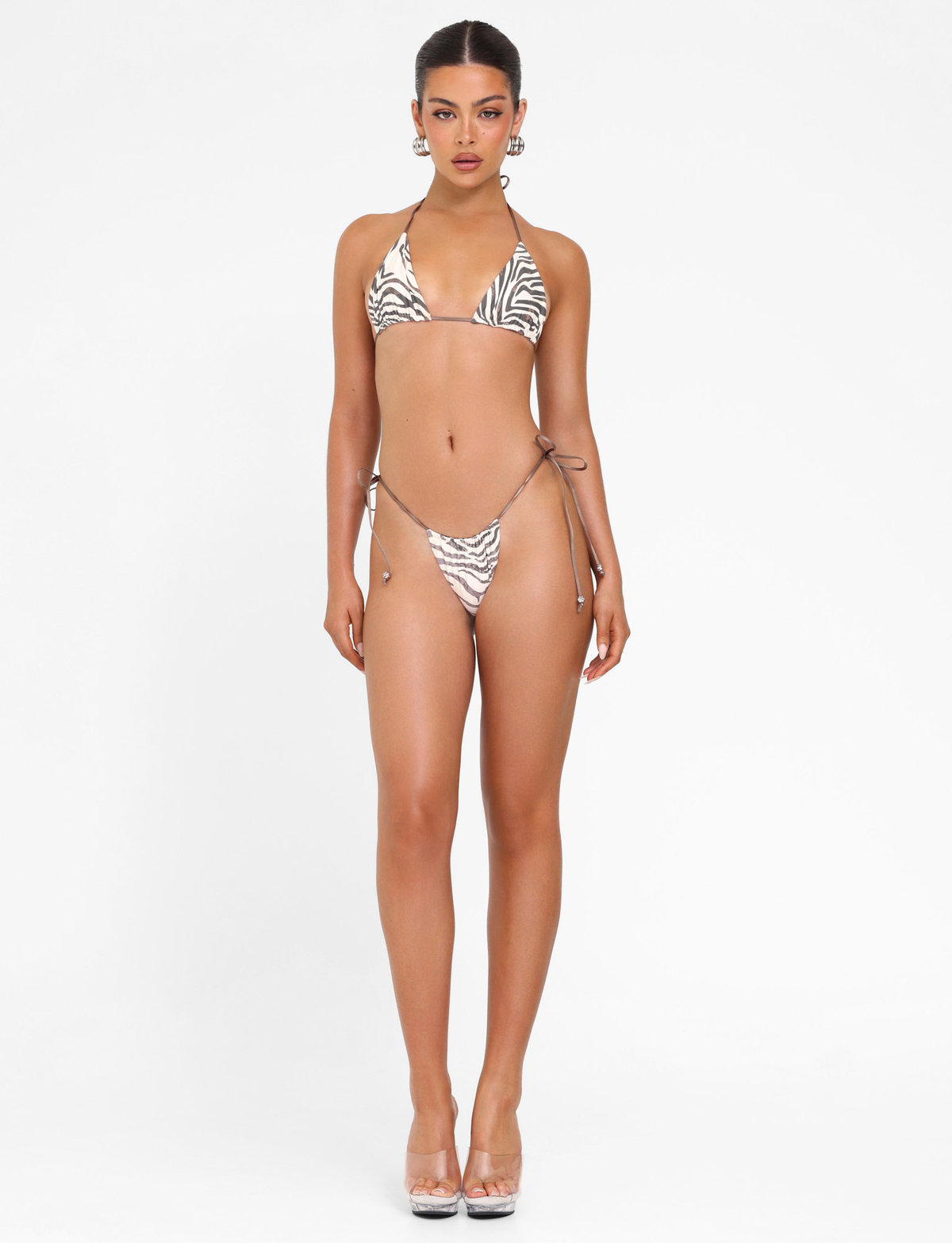 A woman faces forward against a white background, wearing the TIGERMIST AVE BIKINI TOP - NEUTRAL : ZEBRA with triangle cups, paired with clear heels. Her hair is tied back and she accessorizes with large earrings.