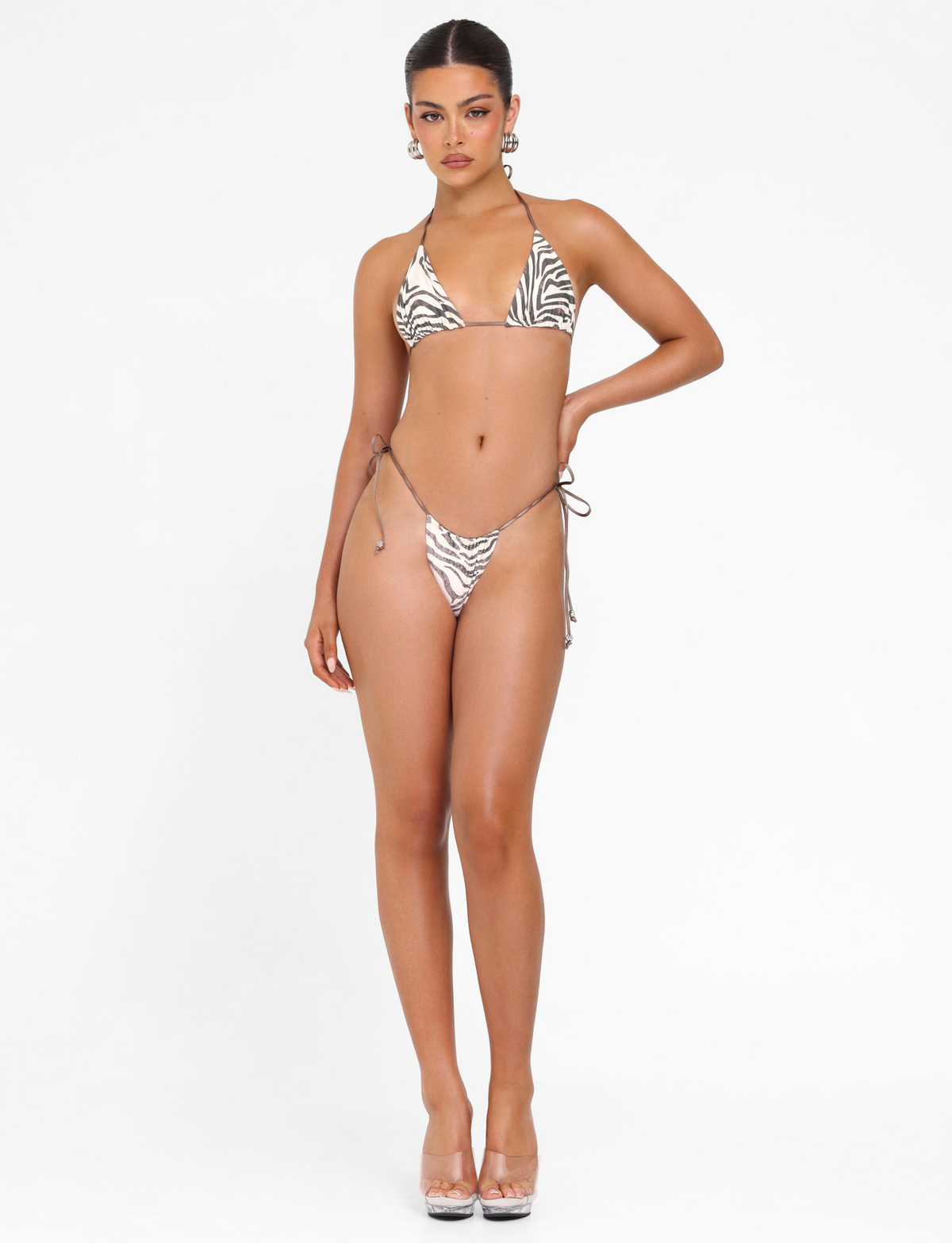 A woman stands against a white background wearing the TIGERMIST AVE BIKINI TOP - NEUTRAL : ZEBRA with triangle cups and clear heels, one hand on her hip and a neutral expression.
