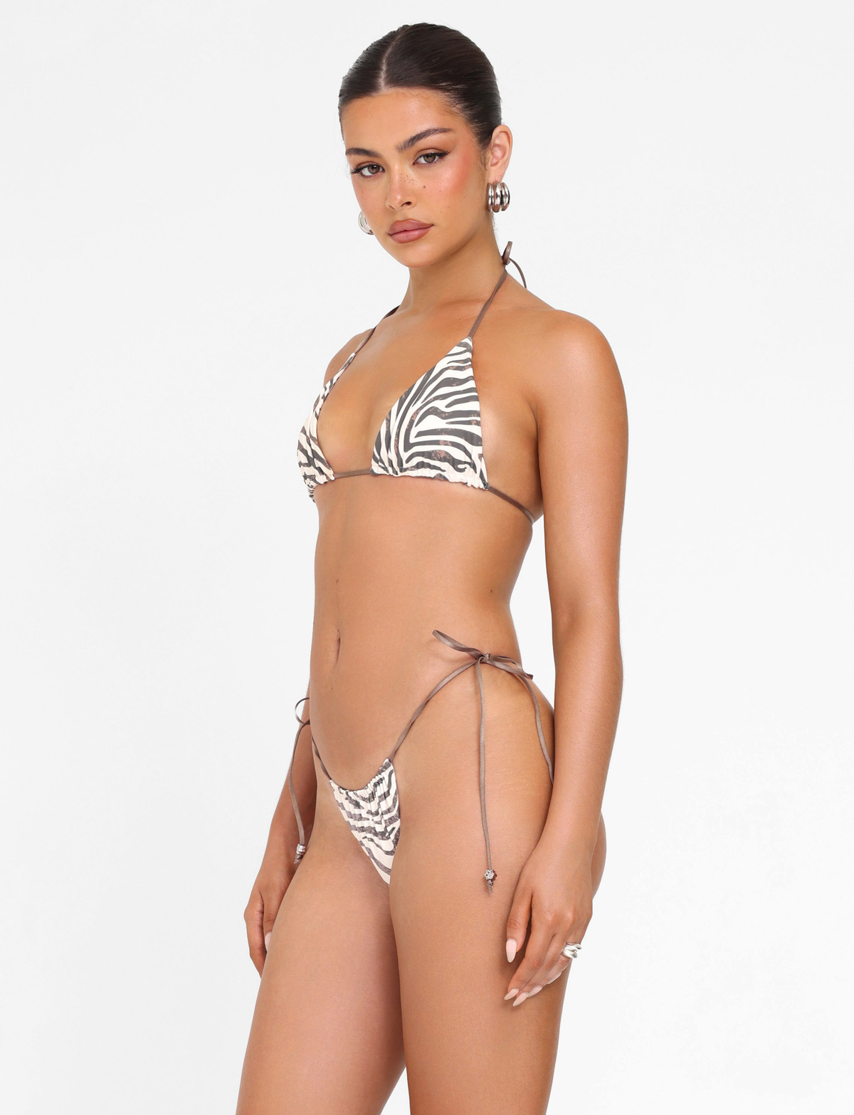 Model wears the TIGERMIST AVE BIKINI TOP - NEUTRAL : ZEBRA, a triangle-cup string bikini, posing sideways with hair pulled back and hoop earrings against a plain white background.